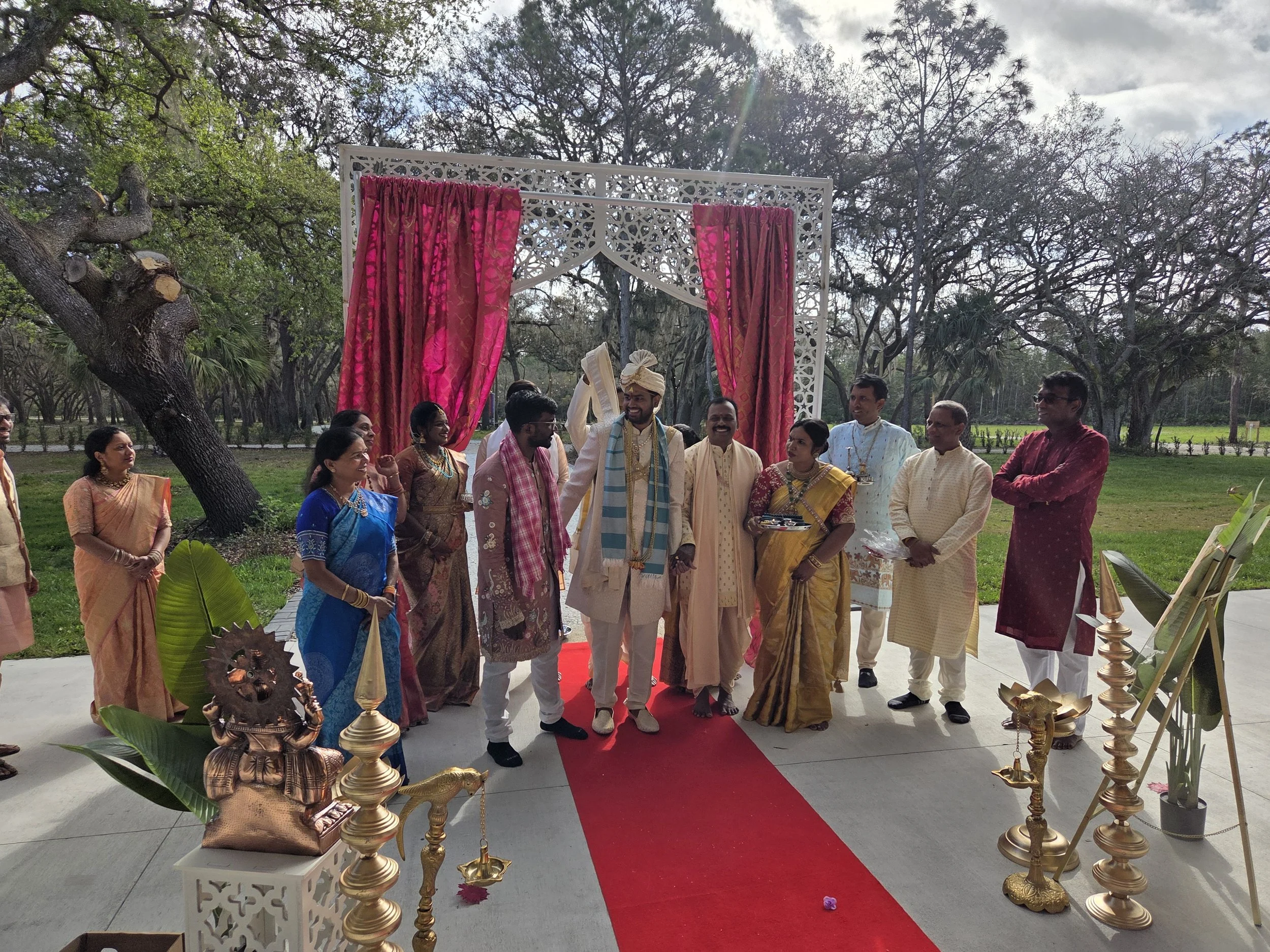 A Hindu wedding ceremony outdoors with a group of people dressed in traditional Indian attire, under a decorative white mandap with pink curtains, on a red carpet, surrounded by trees and greenery.