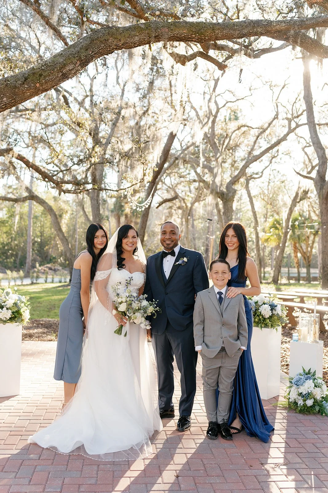 Group of six people at a wedding, standing outdoors in front of trees with sunlight filtering through, decorated with floral arrangements.