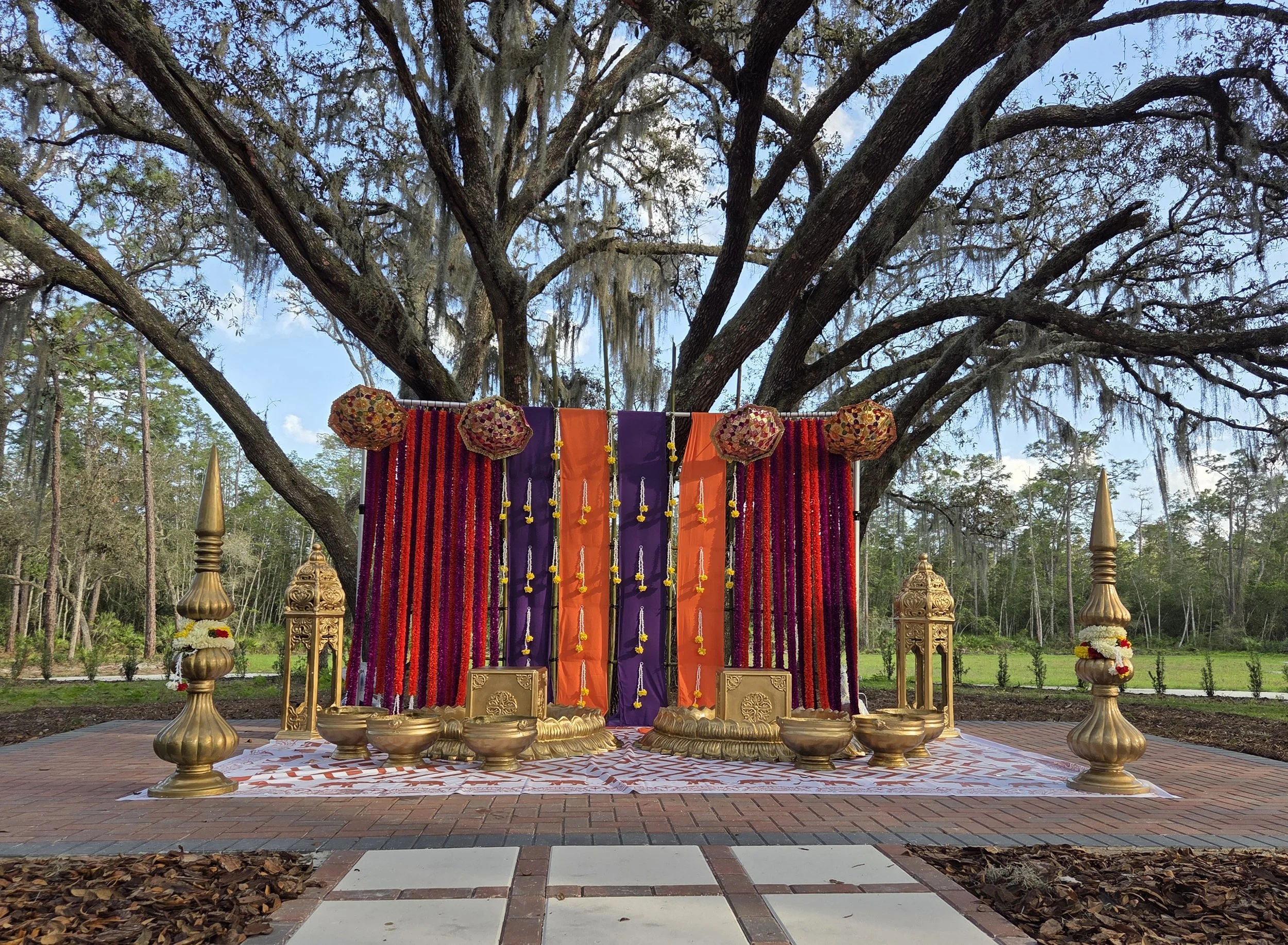 Indian wedding or cultural ceremony setup with gold decorative elements, colorful drapes in purple and orange, hanging floral decorations, and a large tree in the background, outdoors.