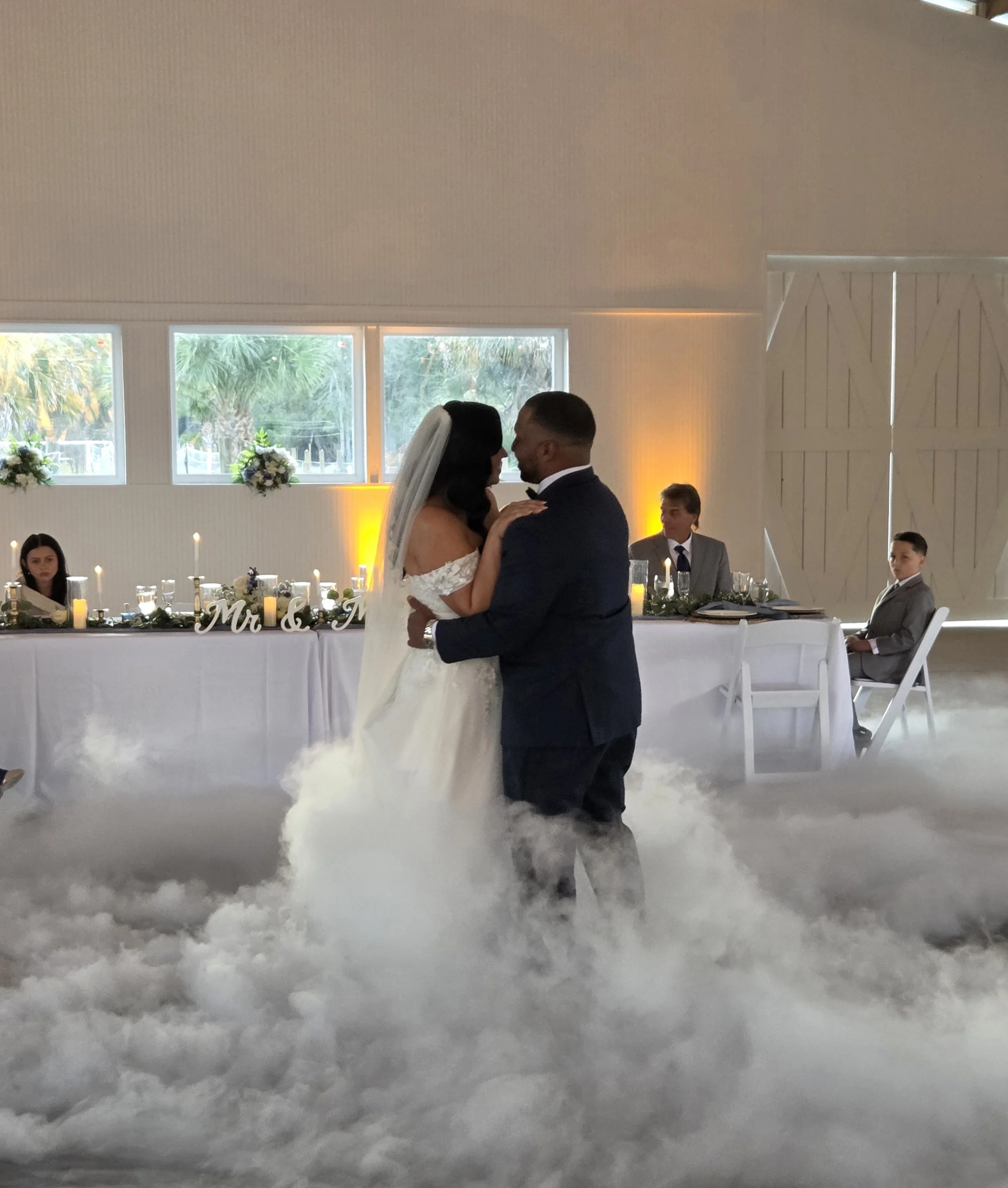 A bride and groom are dancing at their wedding reception, with guests seated at tables decorated with white flowers and candles. The bride is wearing a white wedding gown and veil, and the groom is in a dark suit. The setting is a bright room with la