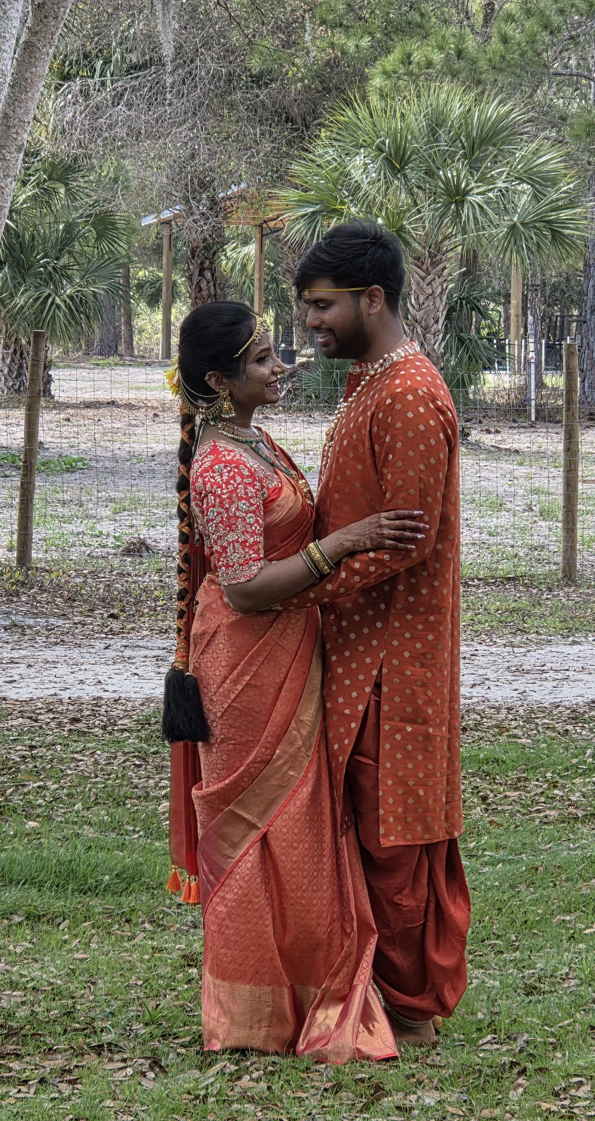 A couple dressed in traditional Indian attire, standing outdoors facing each other and smiling, surrounded by trees and greenery.