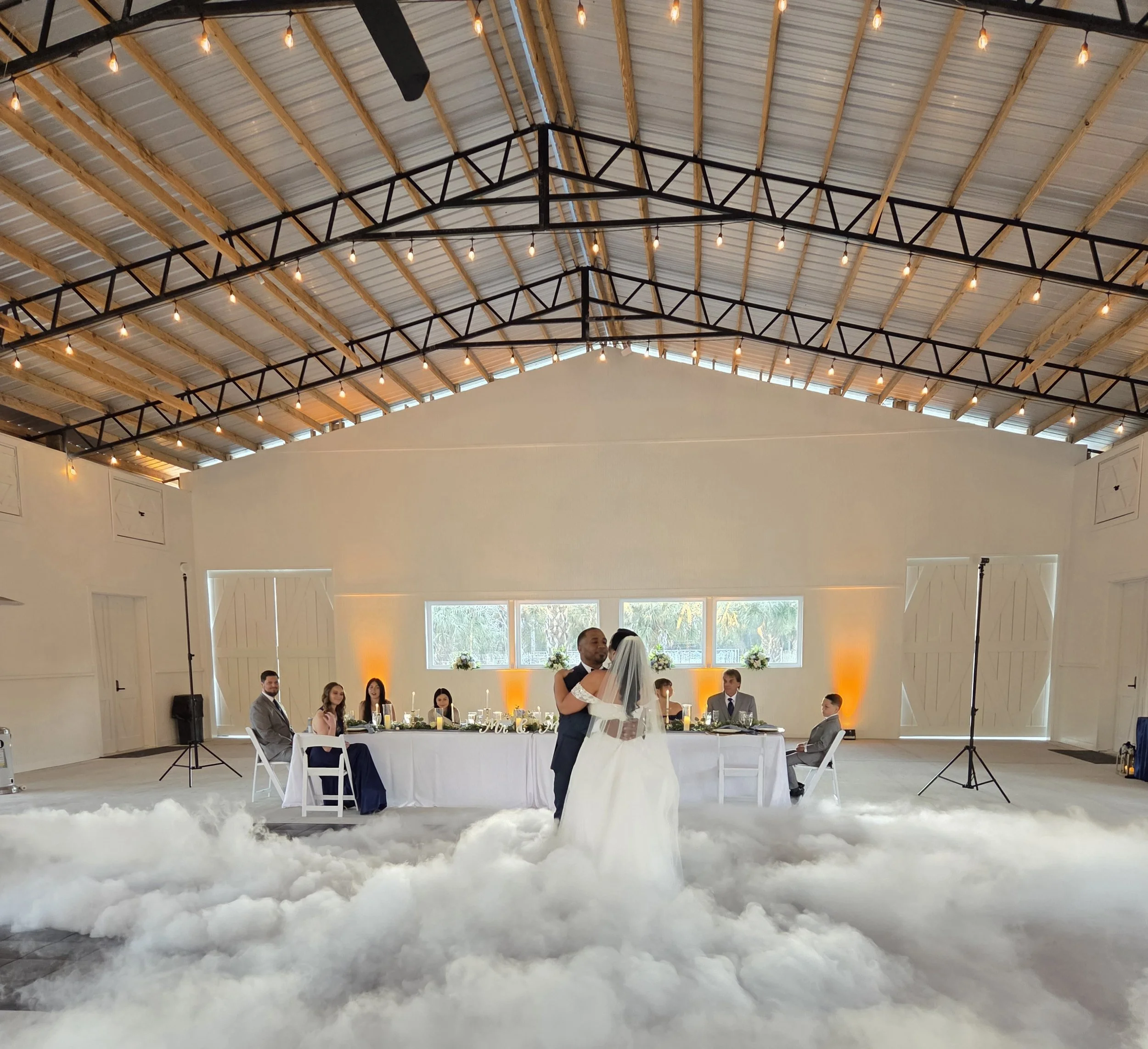 Wedding reception with a bride and groom dancing under a high ceiling, decorated with string lights, with guests seated at a long head table and fluffy white carpet on the floor.