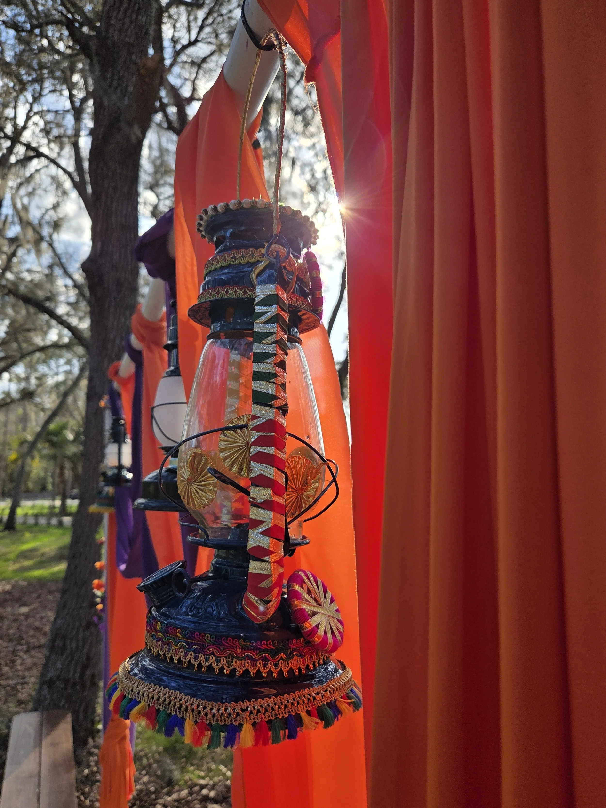 Colorful lanterns hanging on a string with orange fabric backdrop and trees in the background, sunlight visible through the fabric.