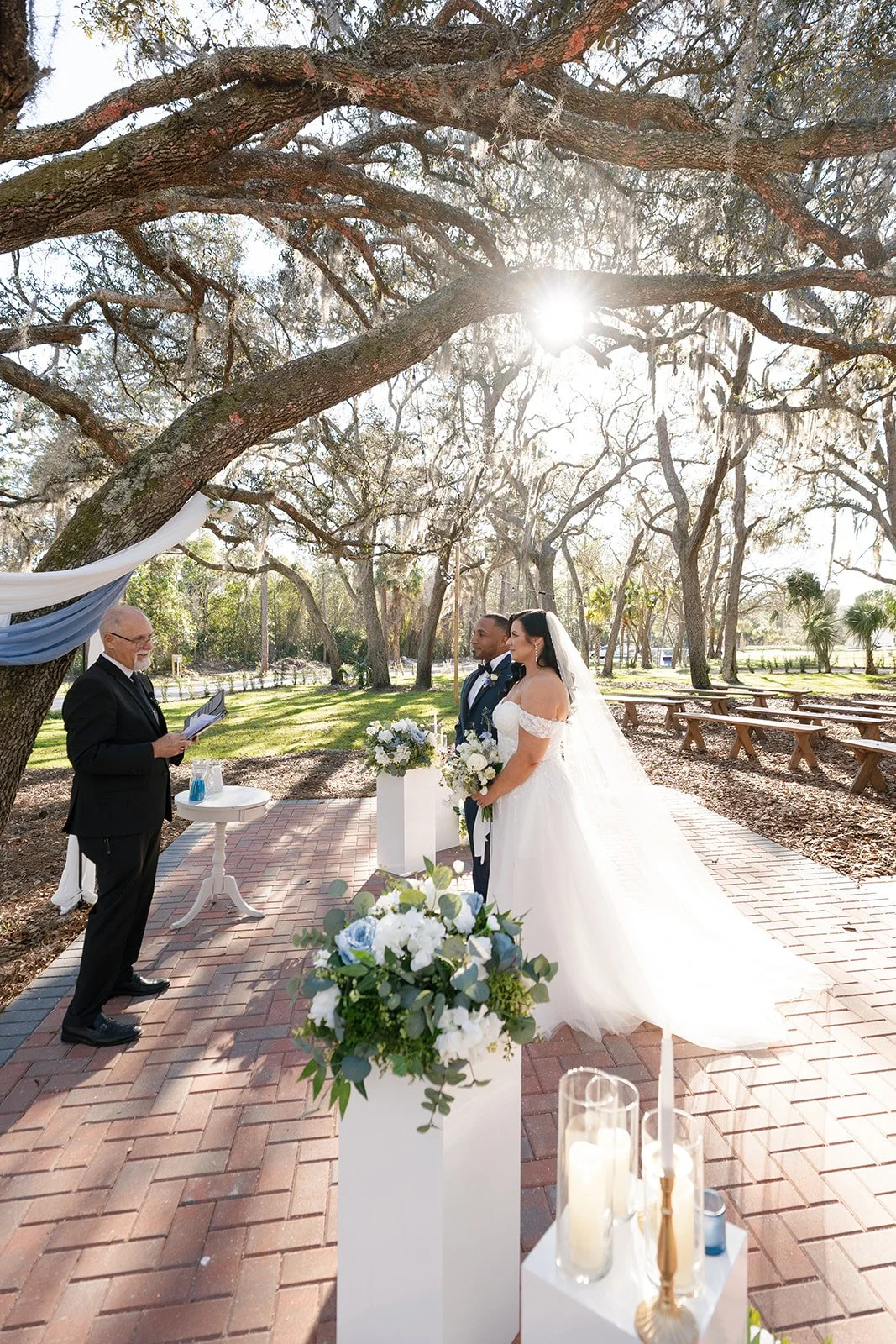 A wedding ceremony outdoors under a large tree with sunlight shining through. The bride and groom stand facing an officiant. The bride wears a white off-shoulder wedding gown and holds a bouquet of flowers. The groom wears a dark suit with a bow tie.