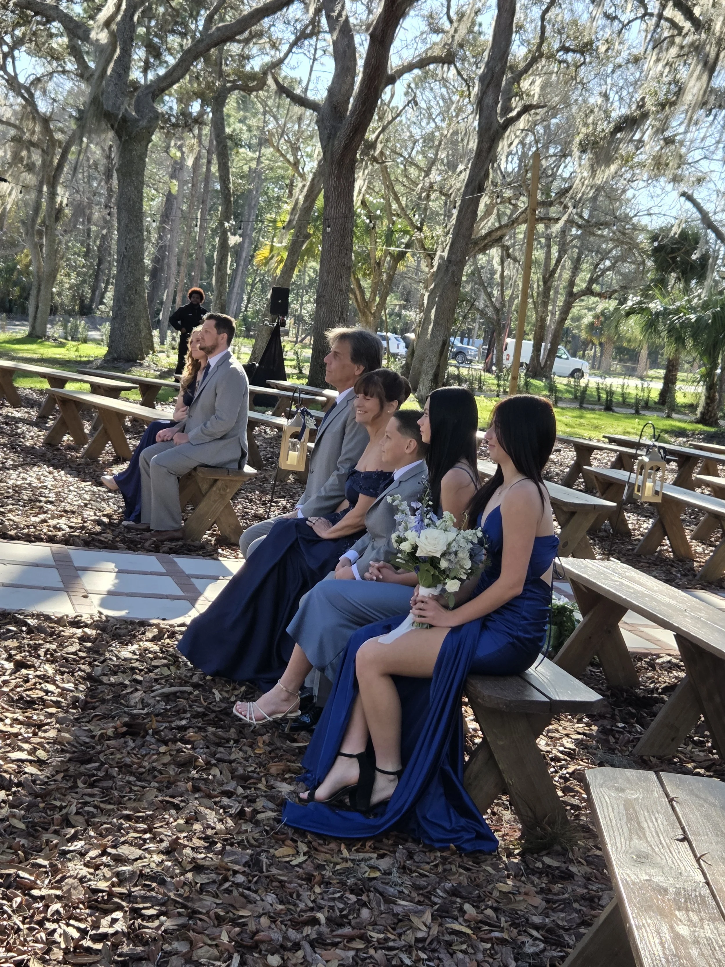 People attending an outdoor wedding ceremony seated on wooden benches amid tall trees, with sunlight filtering through the branches.