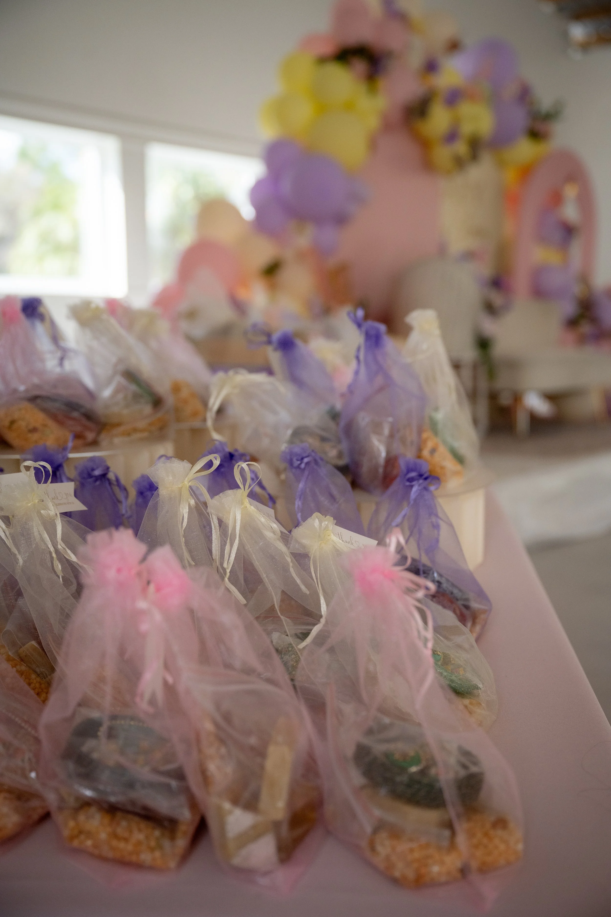 Clear treat bags with purple and pink ribbons, filled with colorful candies, arranged on a table for a celebration, with a pastel-colored cake in the background.