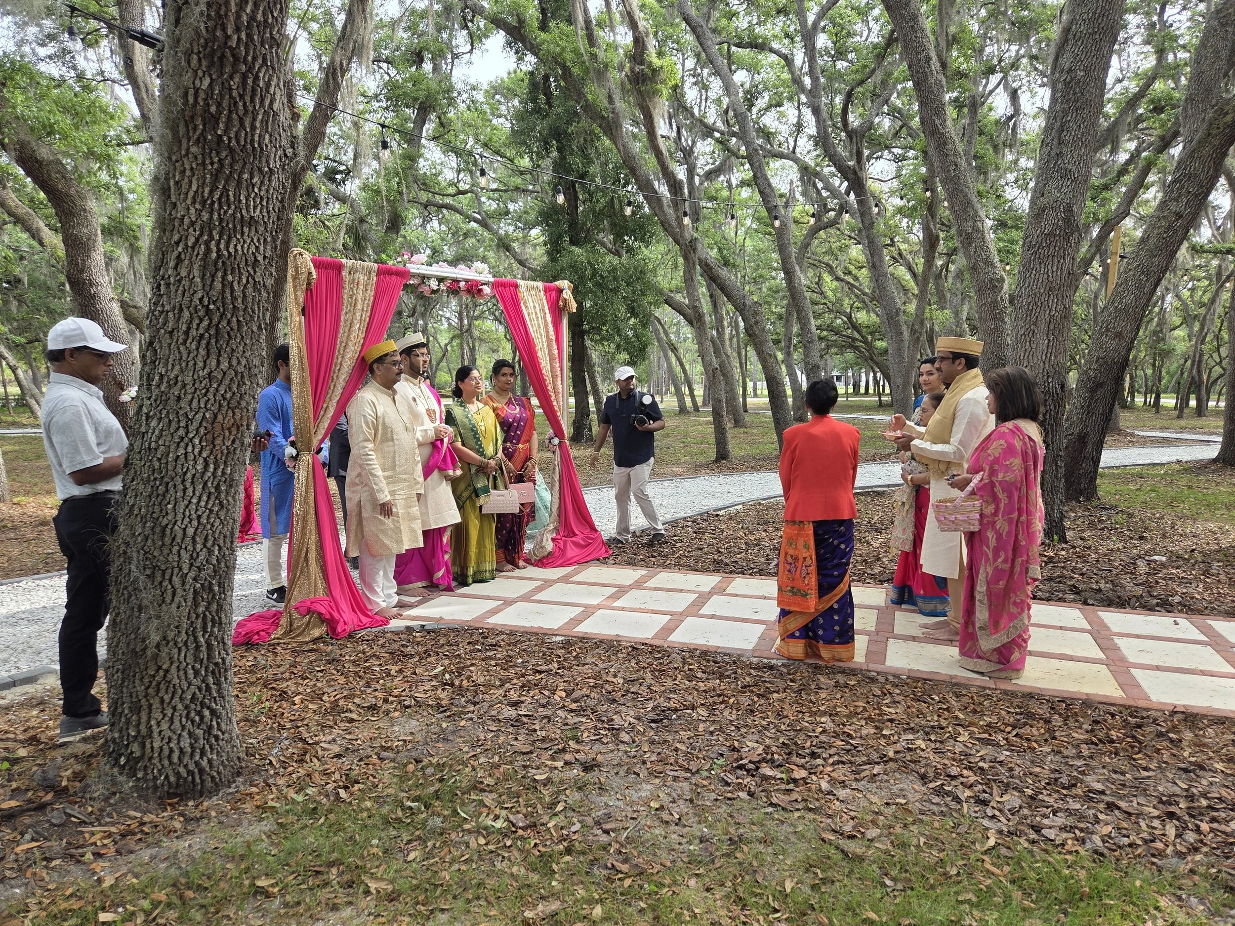 A group of people, some in traditional Indian attire, gather outdoors under tall trees for a ceremony marked by a decorated pink and gold backdrop with flowers. There are multiple men and women, some wearing turbans, and a photographer capturing the 