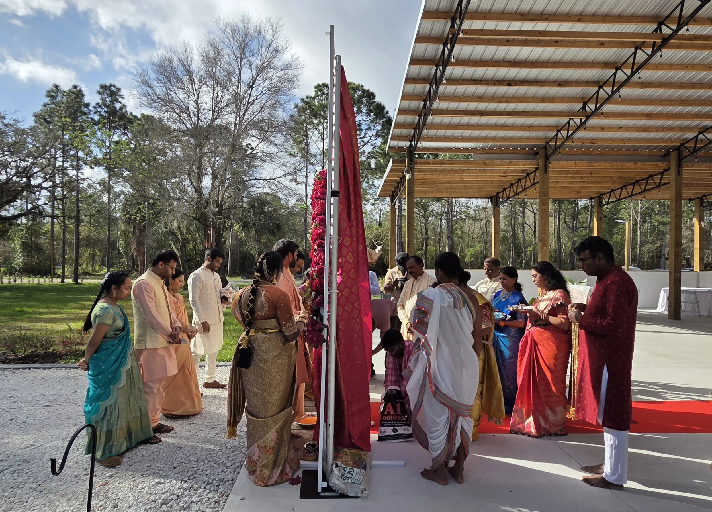 A group of people dressed in traditional Indian clothing participating in a ceremony outdoors under a wooden pavilion, with trees and cloudy sky in the background.