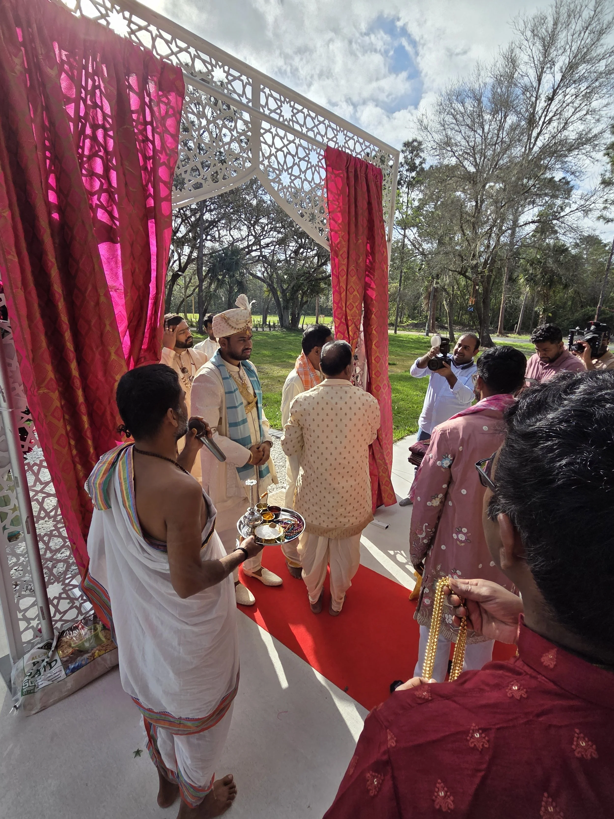 A Hindu wedding ceremony outdoors with a group of men dressed in traditional attire. One man is holding a tray with ceremonial items, while others are gathered in front of a decorated arch with pink curtains.