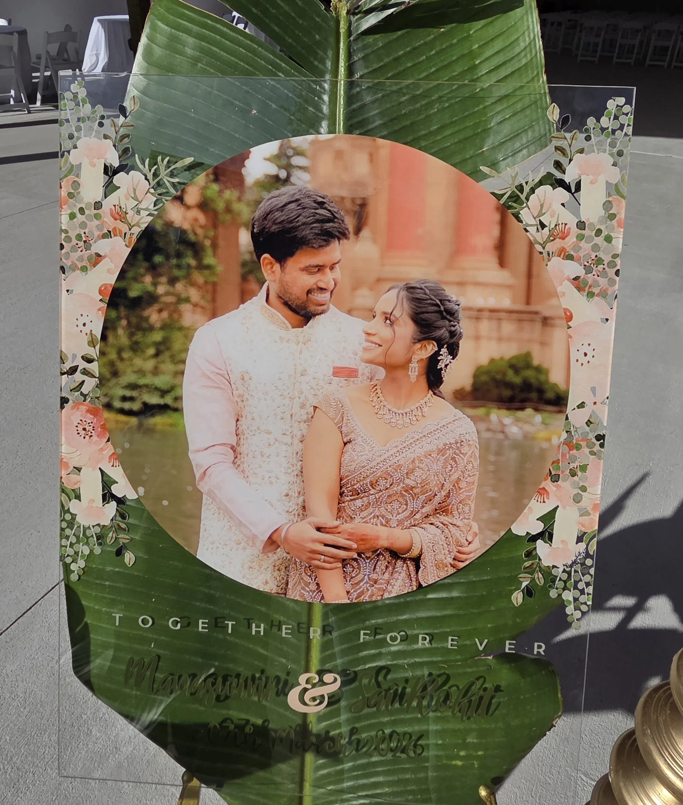 A wedding photo of a groom and bride, smiling and looking at each other, with lush greenery and trees in the background. The photo is overlaid on a large green leaf. Over the image, there is decorative floral border and text that reads, 'TOGETHER FOR