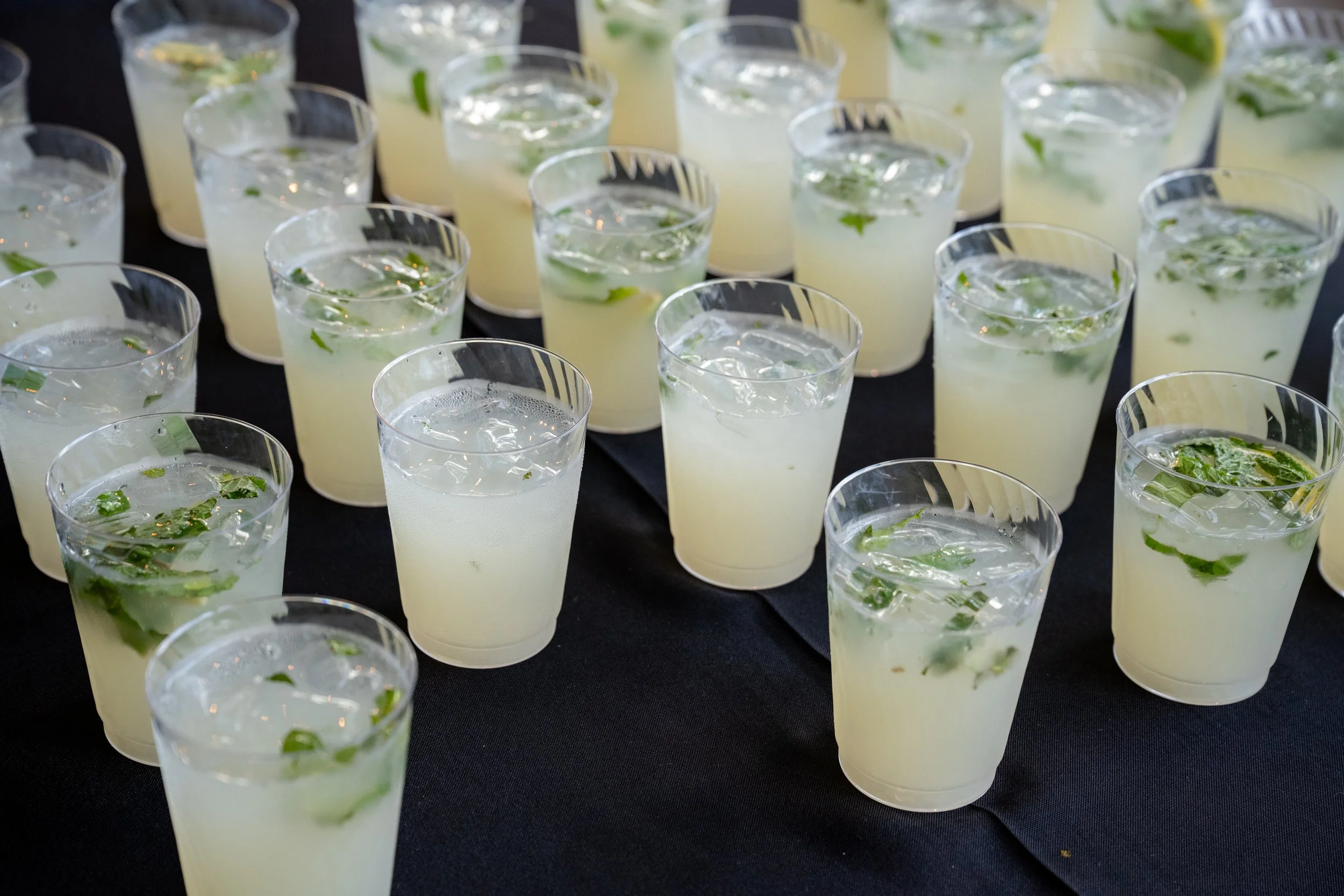 Multiple clear plastic cups filled with a light yellow beverage, ice, and fresh mint leaves, arranged on a dark surface.