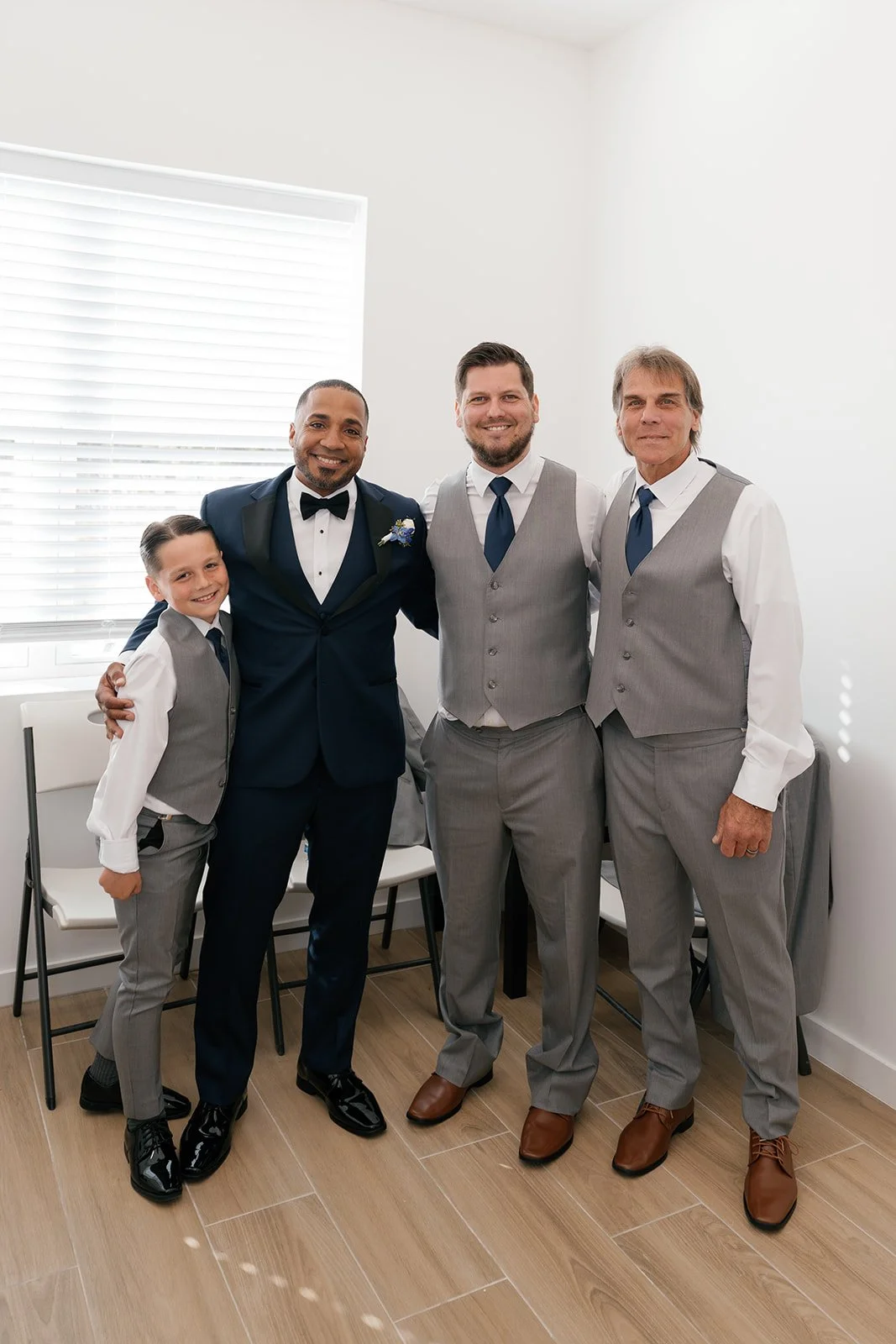 Group photo of five men dressed in formal suits, including a groom in a navy tuxedo with a bow tie, and four men in grey vests and trousers, with one young boy also in a grey vest, all smiling indoors near a window with white blinds.