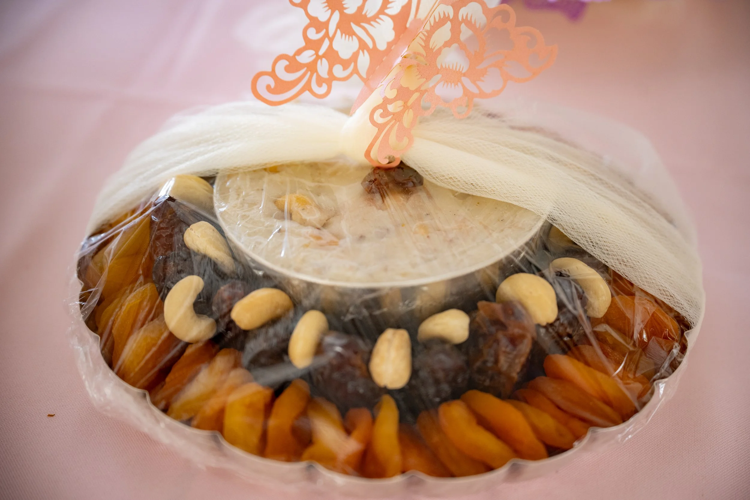 A gift wrapped in clear plastic with a pink decorative paper flower on top, containing various dried fruits and nuts.