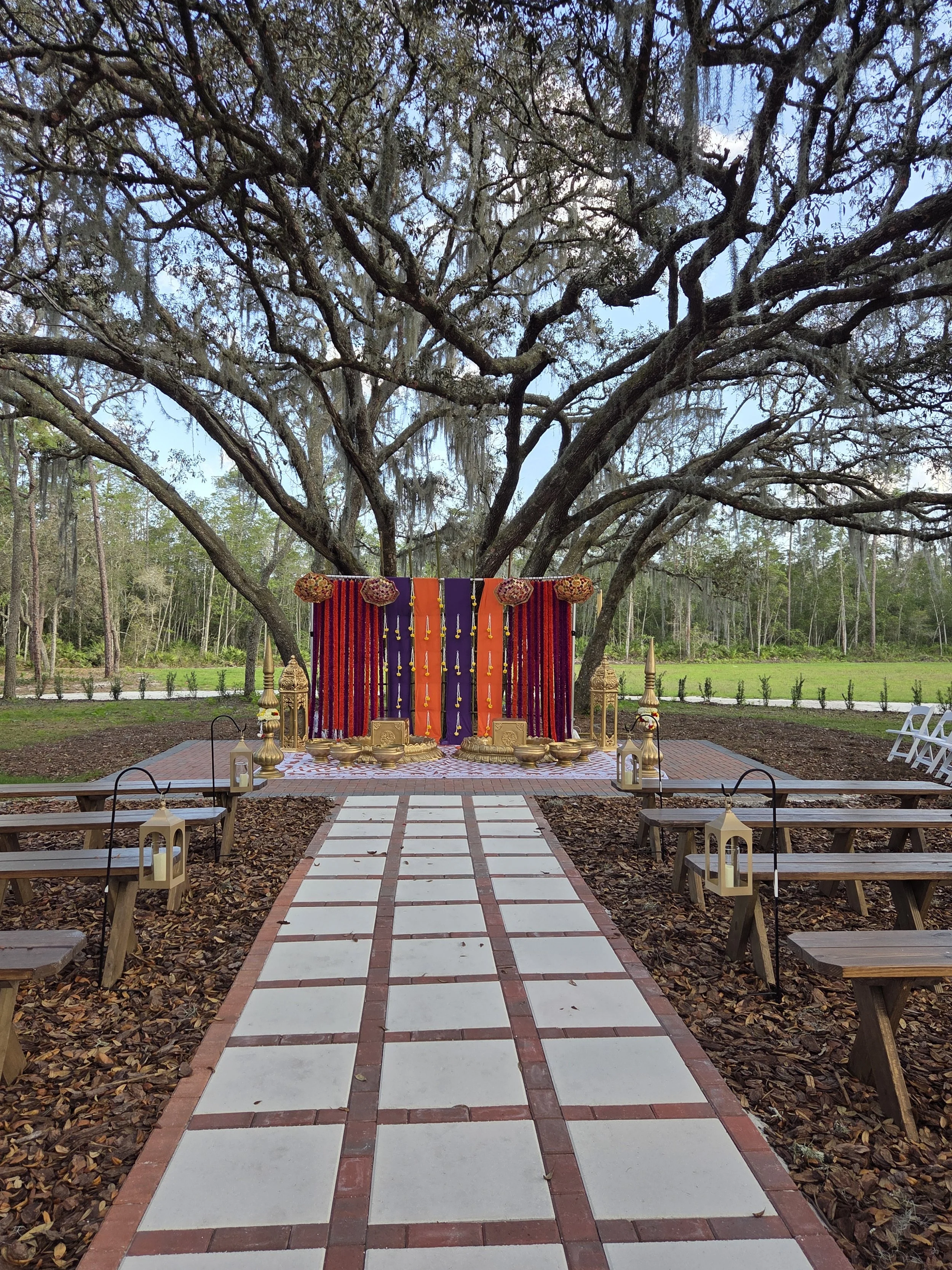 A decorated altar setup outdoors under a large tree with spreading branches, surrounded by benches and lanterns, possibly for a wedding or special ceremony.