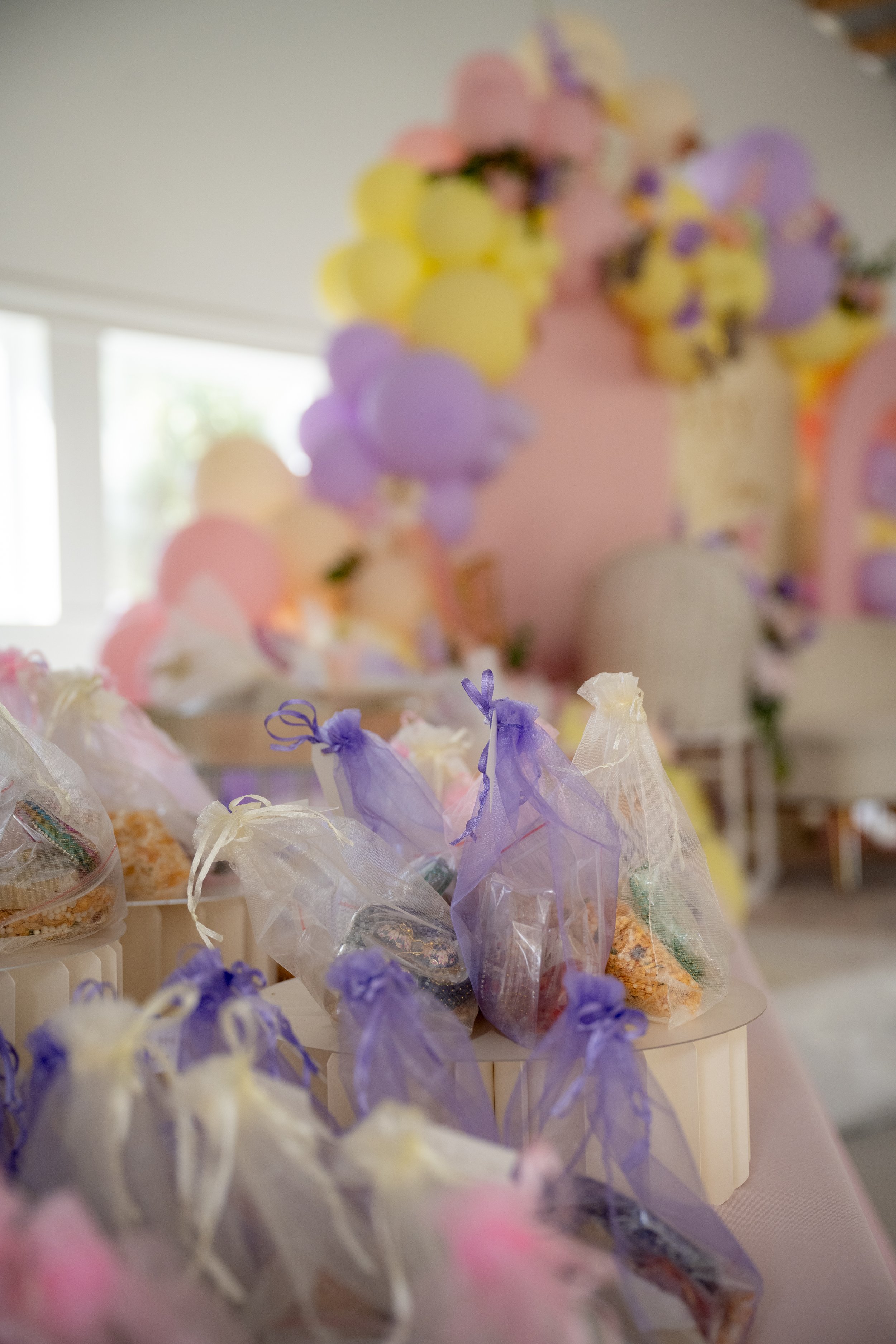 Colorful candies wrapped in pink, purple, and white tissue paper, arranged on a table with a blurred pastel-colored balloon and decoration in the background.
