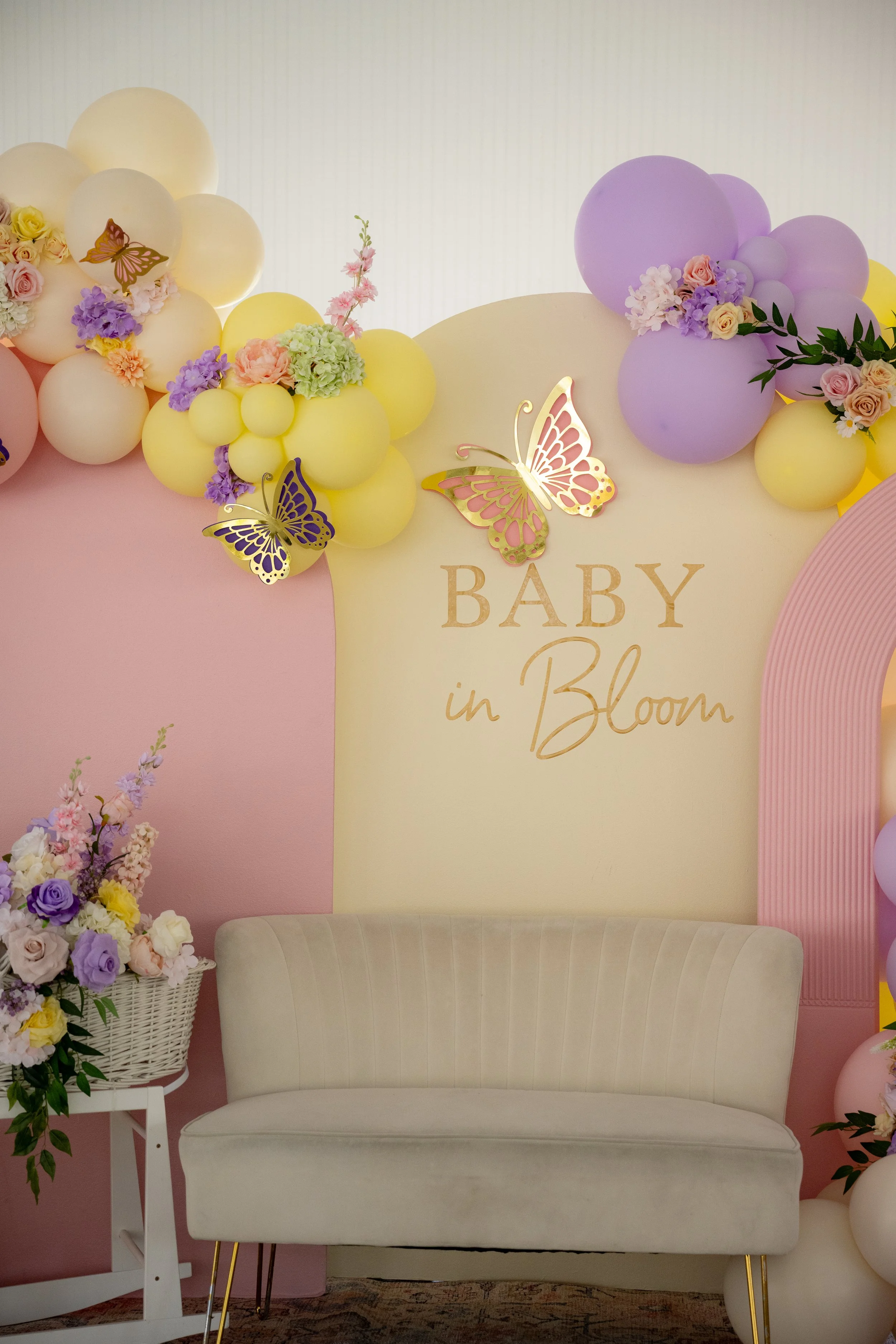 Decorative backdrop with pastel balloons, flowers, and butterfly cutouts, with a beige sofa and floral arrangements in baskets, featuring the words 'Baby in Bloom'.