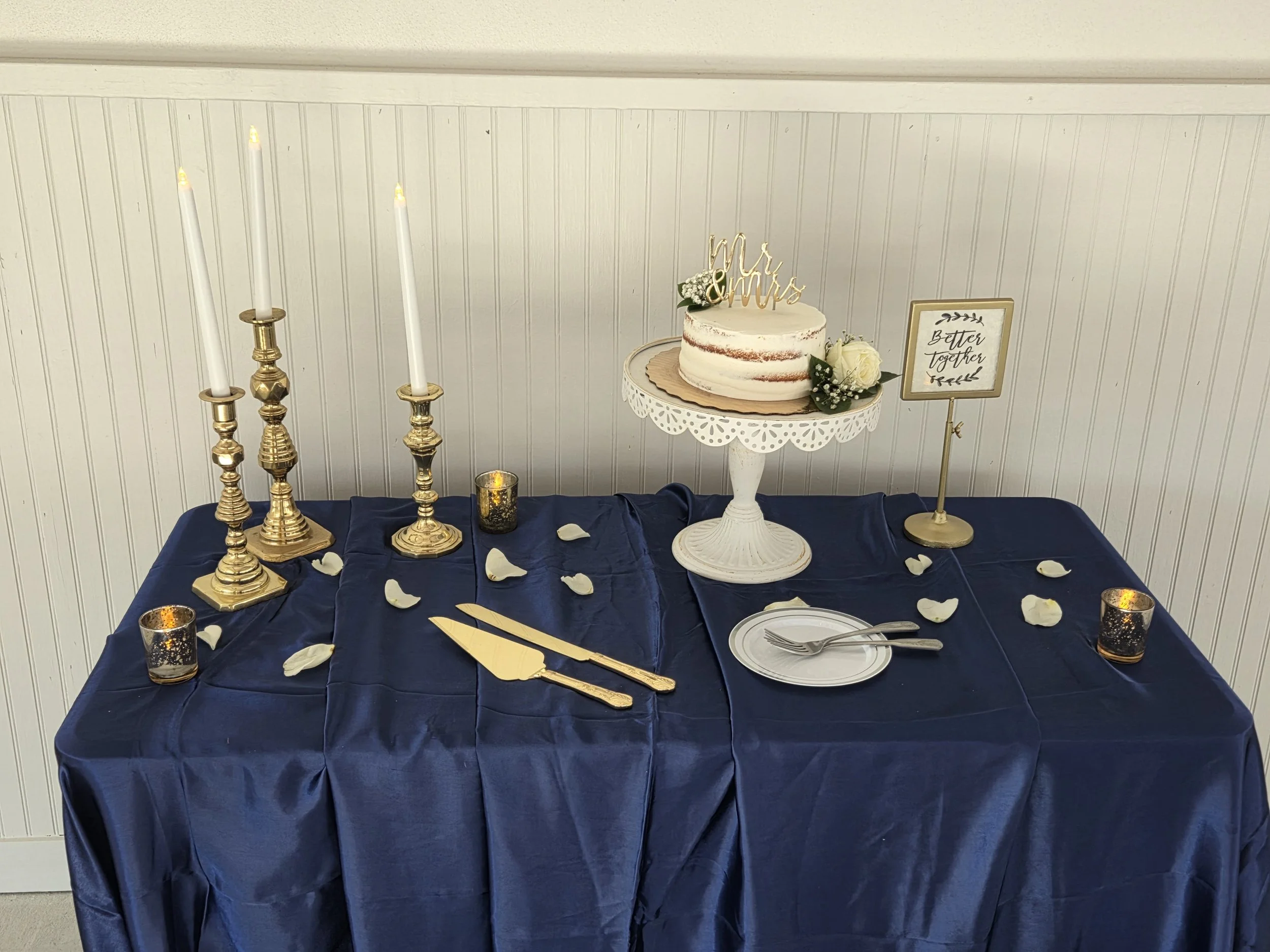 A wedding dessert table featuring a semi-naked wedding cake with a 'Mr. & Mrs.' topper, surrounded by flowers, candles, and gold cutlery, with a sign that reads 'Better together.'