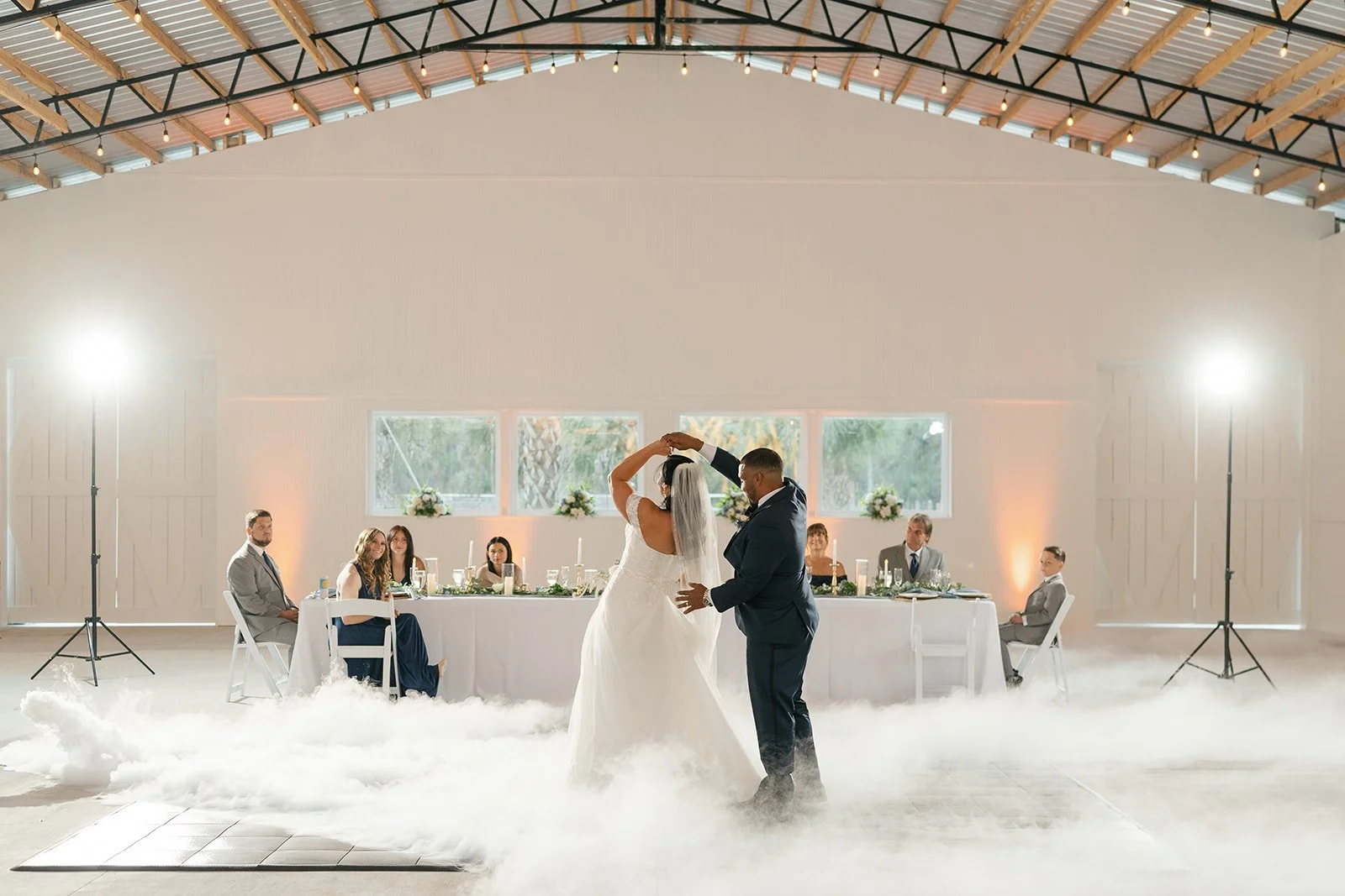 A bride and groom dance at their wedding reception with guests sitting at a long table behind them. The space is decorated with flowers and candles, with fog on the floor and bright lighting.