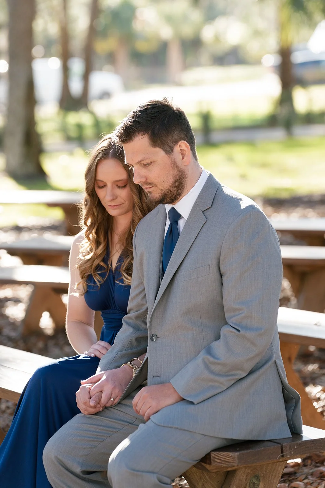 A man and woman sitting closely together on a park bench during daytime, with their heads bowed and holding hands, dressed in formal attire.