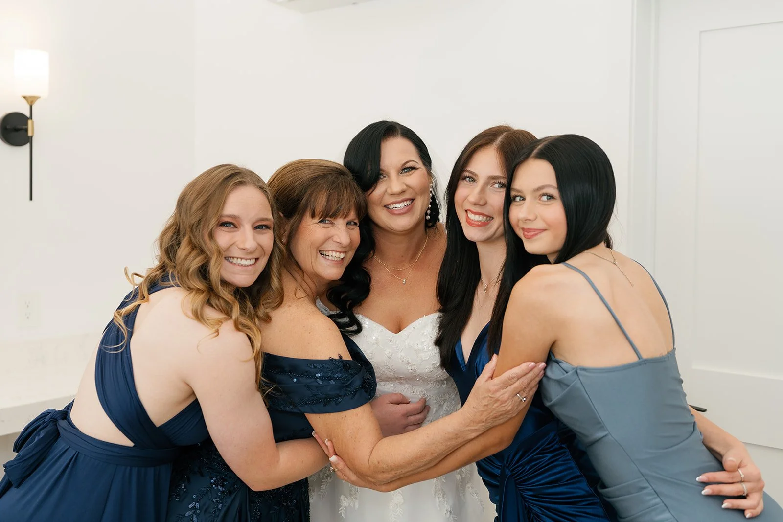 Five women in formal dresses smiling and hugging each other indoors, with a white wall in the background.