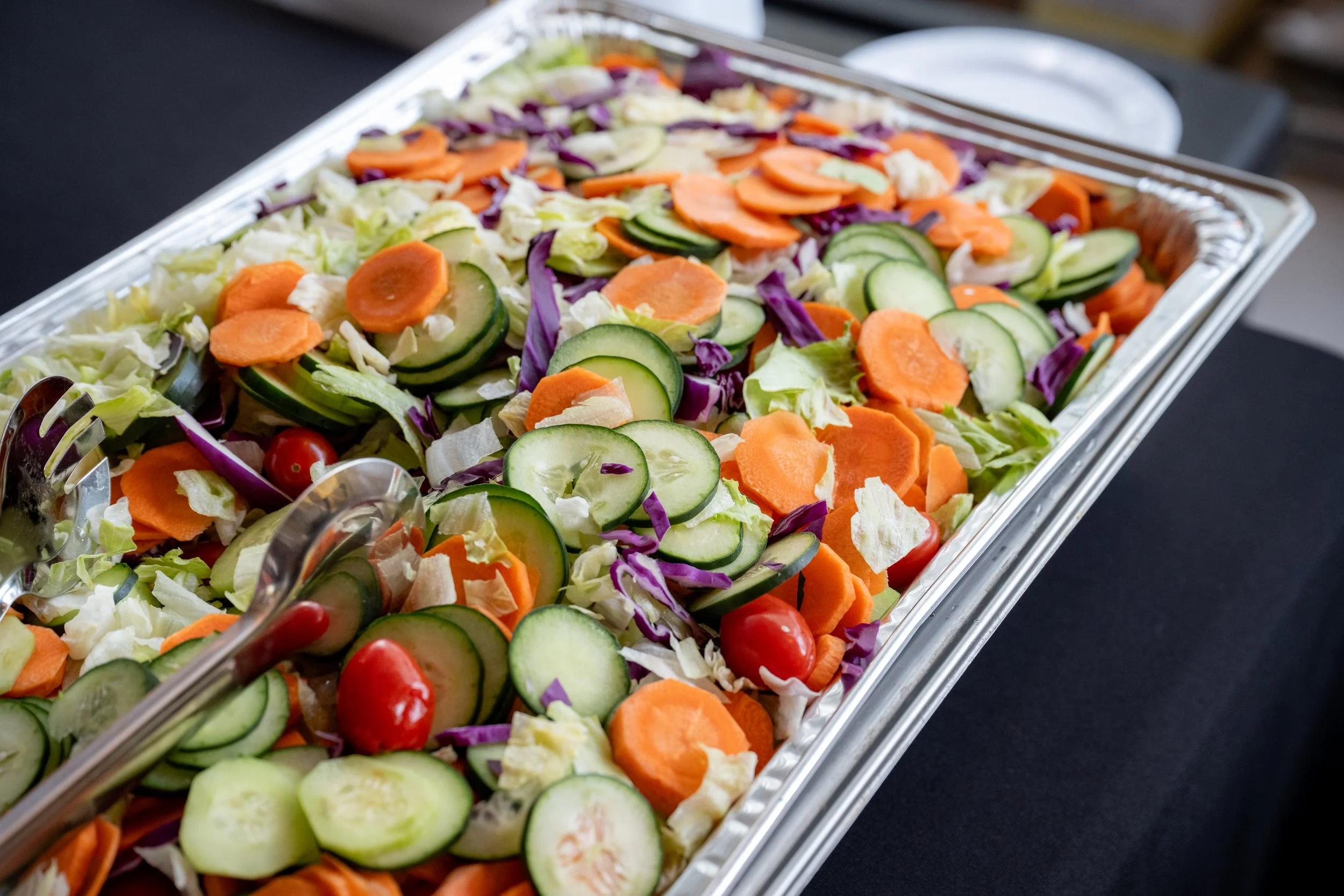 A large tray of fresh salad with sliced cucumbers, shredded purple cabbage, chopped lettuce, sliced carrots, cherry tomatoes, and some white cheese.