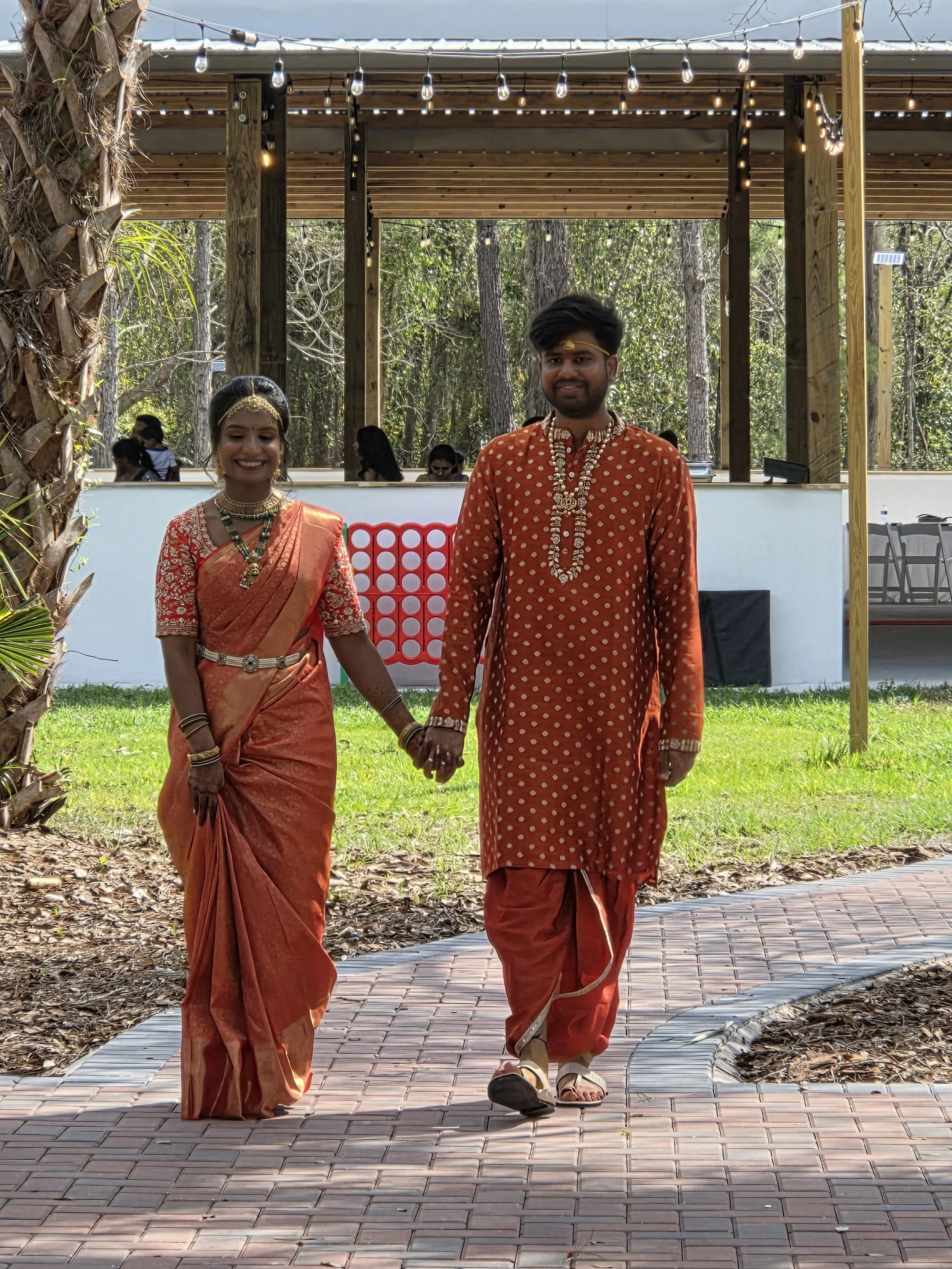 A young couple dressed in traditional Indian clothing, holding hands and walking outdoors on a curved brick path surrounded by greenery and tall trees. The woman is smiling and wearing a red and gold saree with jewelry, while the man is wearing an or