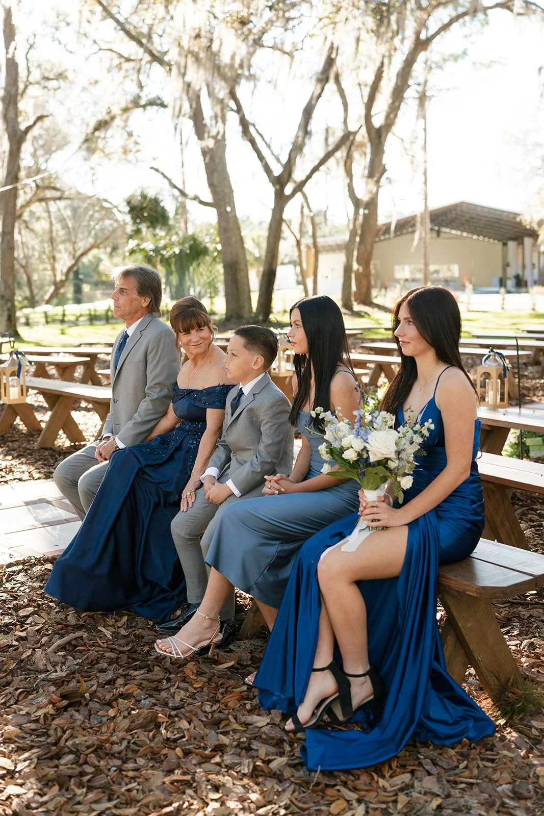 People sitting outdoors during a wedding ceremony surrounded by trees with sunlight shining through.