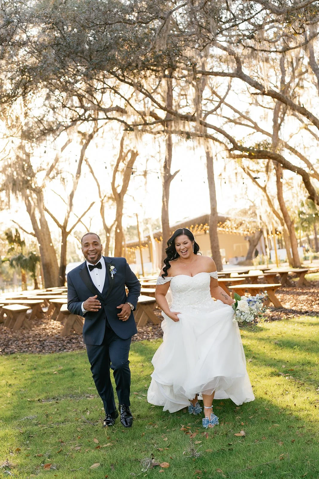 A bride and groom walking outdoors in a park on their wedding day, smiling and holding flowers, with large trees and picnic tables in the background during sunset.