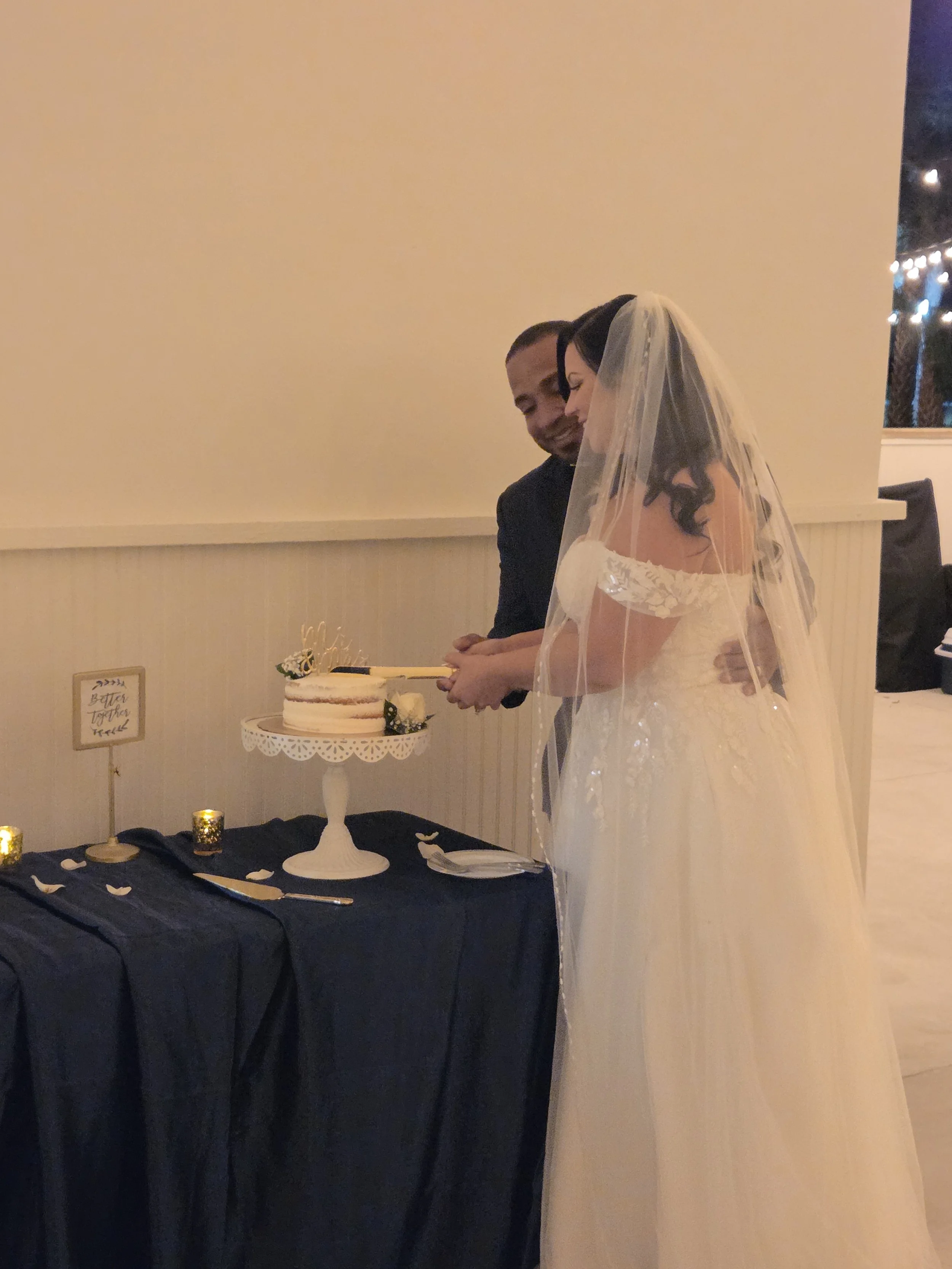 A bride and groom during their wedding ceremony, with a wedding cake on a stand, a sign that says "Better Together," and decorative candles on a table