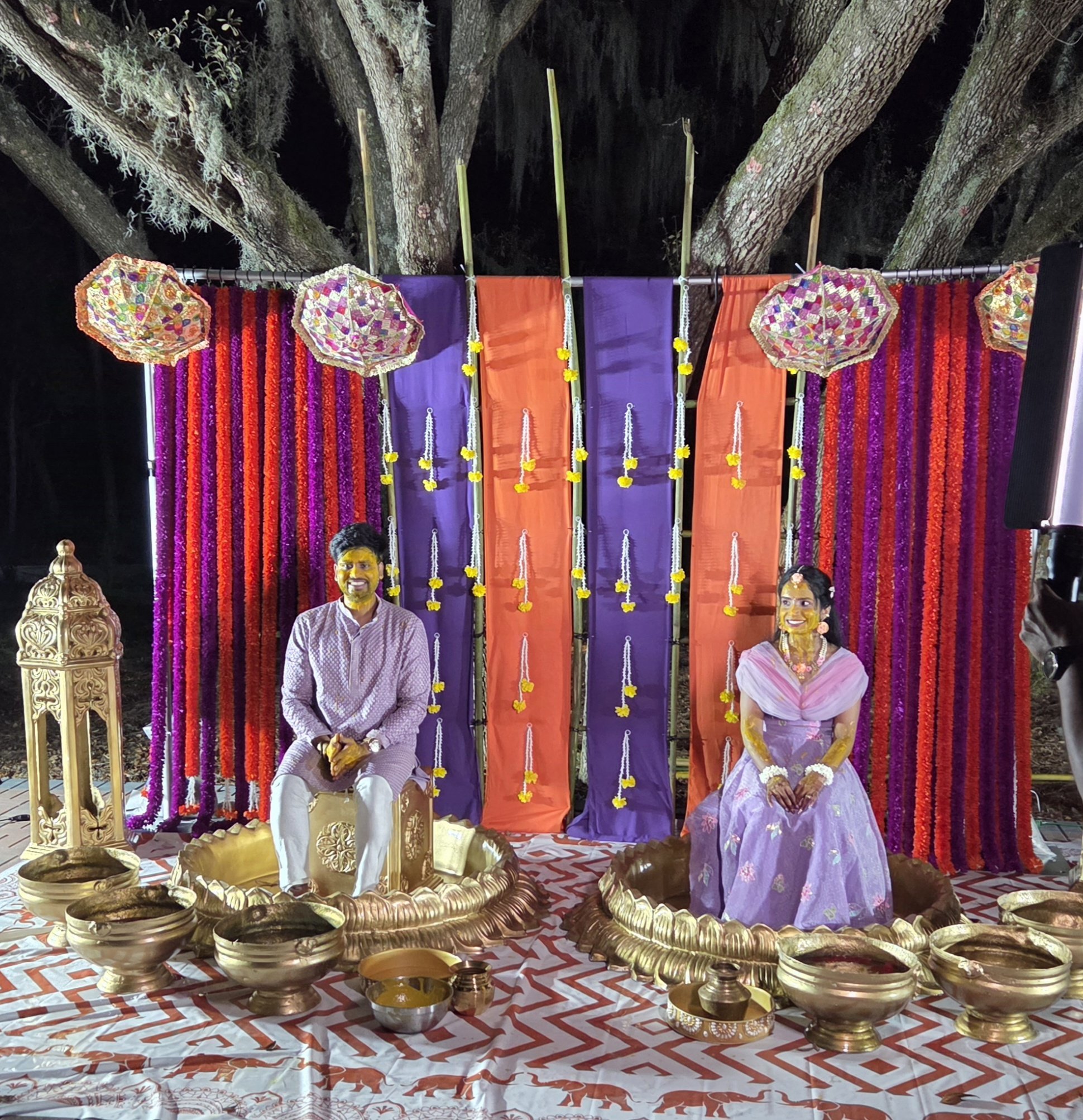 A cultural or religious ceremony setup with a backdrop of hanging colorful fabrics in purple, orange, and pink. Traditional brass or metal bowls are placed at the front, and intricately carved wooden decorations are on the sides. Statues of deities o