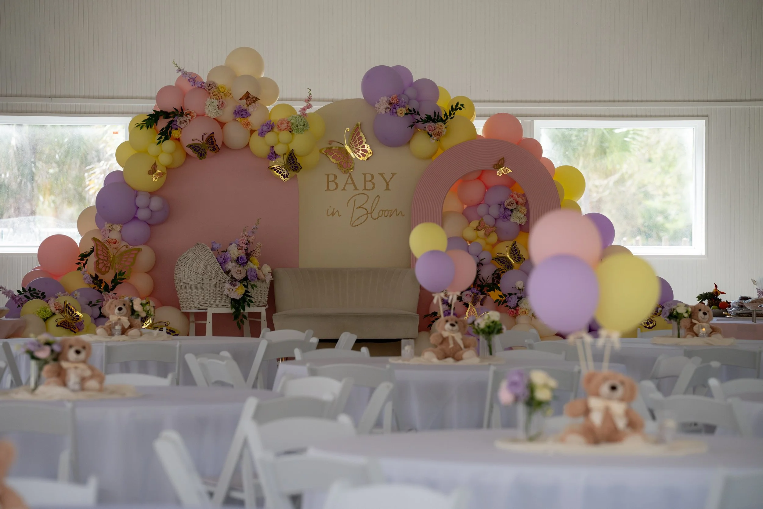 Decorative backdrop with balloons, flowers, and butterfly cutouts at a baby shower event, featuring a pink and purple color scheme with a sign that says 'Baby in Bloom'.