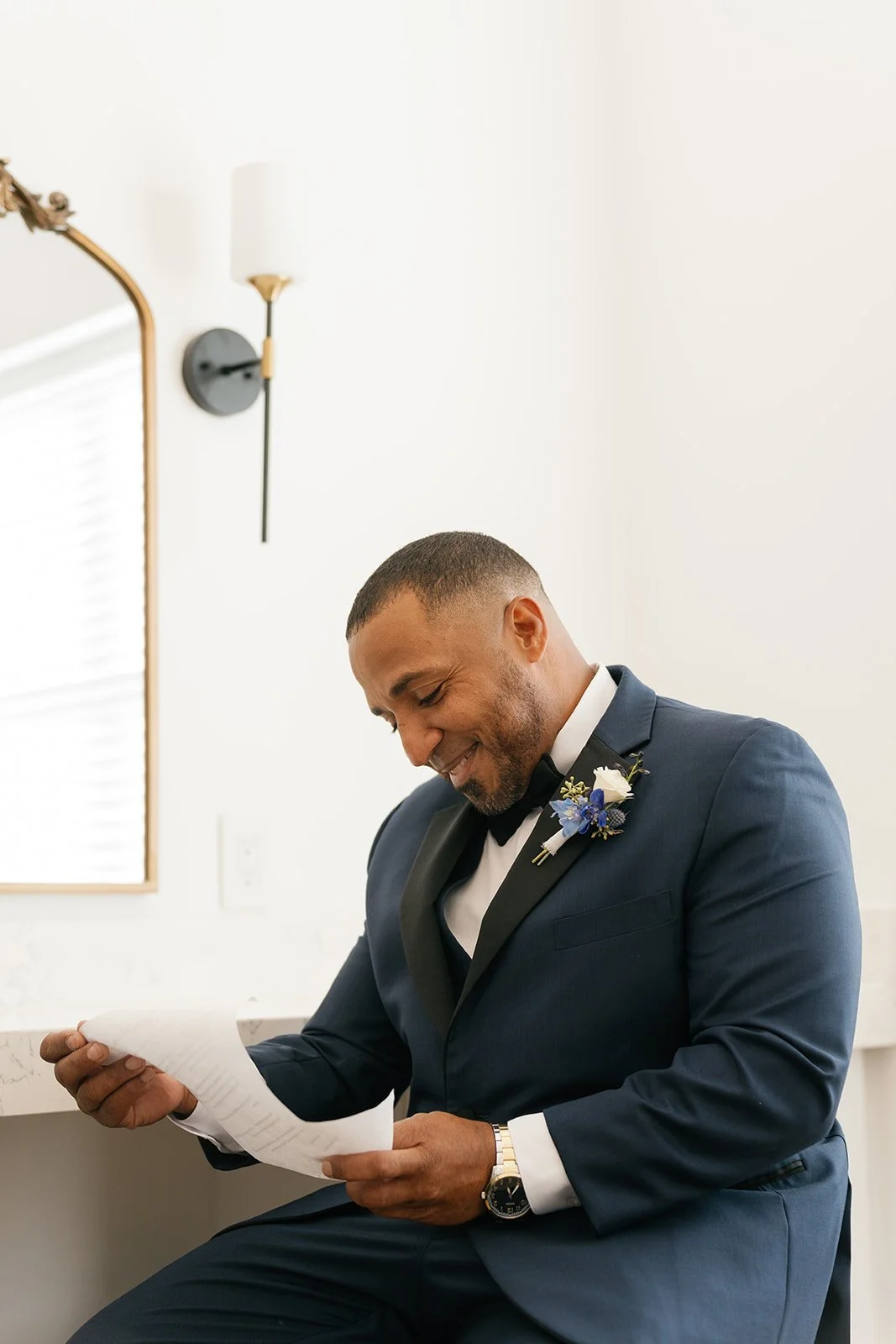A man in a navy tuxedo with a boutonnière, sitting and smiling while reading a piece of paper in a well-lit room with minimal decor.