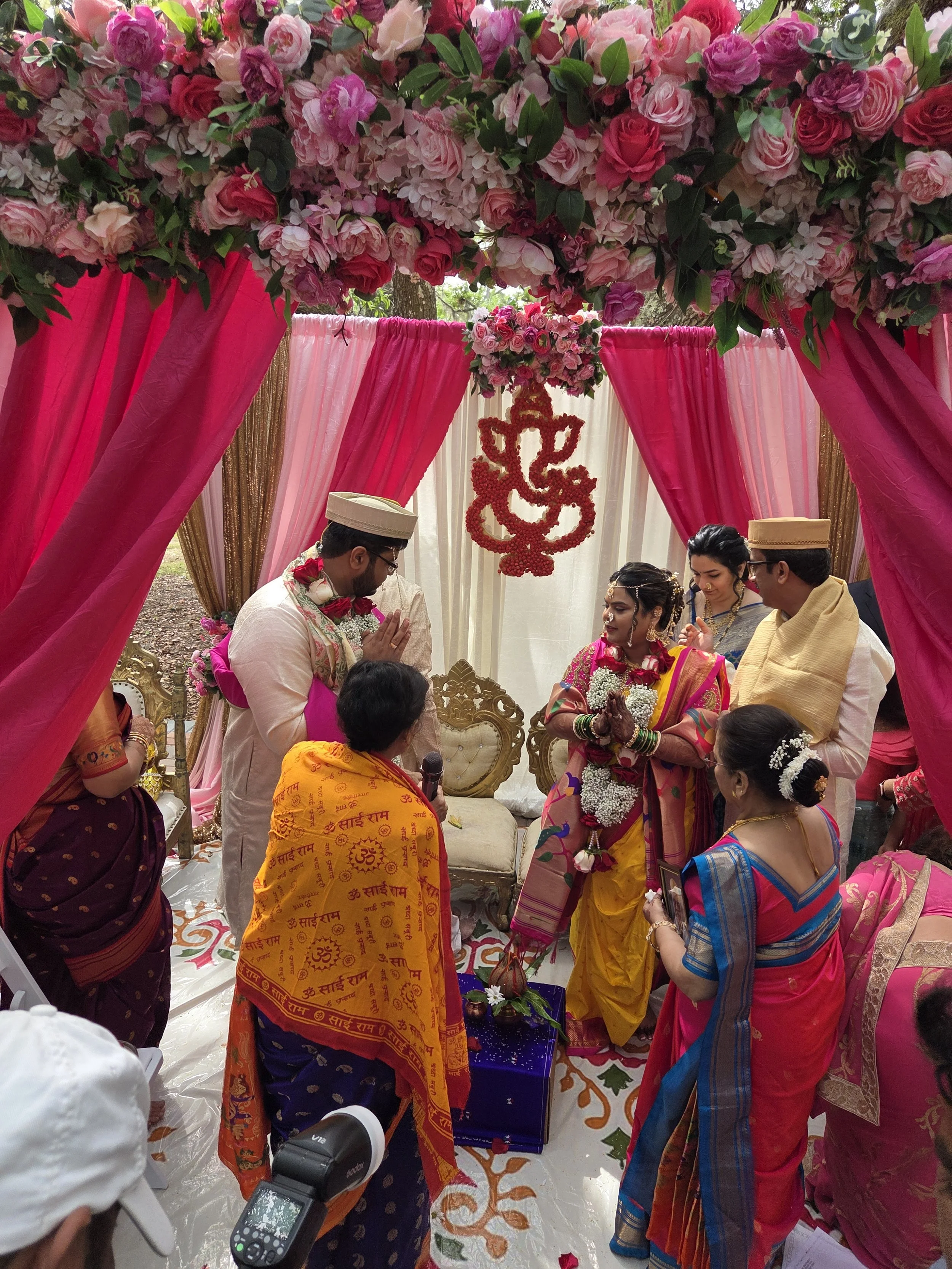 Hindu wedding ceremony with multiple women and men dressed in traditional colorful attire, under a floral canopy decorated with pink and red roses, and a hanging red flower design.