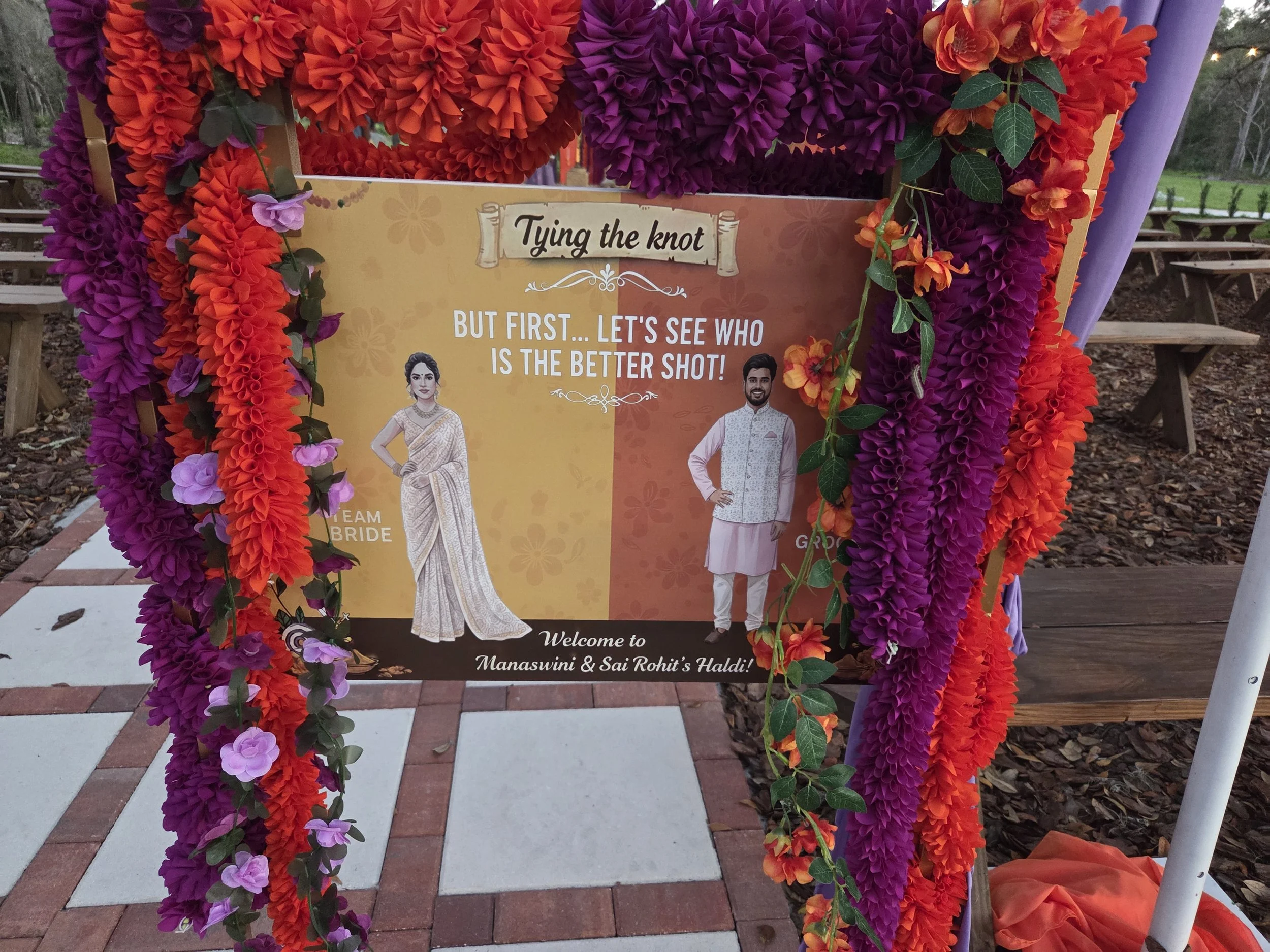 A decorative board with orange and purple paper flowers and green leaves, celebrating a wedding or Haldi ceremony for Manaswini and Sai Rohit, with photos of a woman in traditional attire and a man in traditional Indian clothing, on a pathway with be