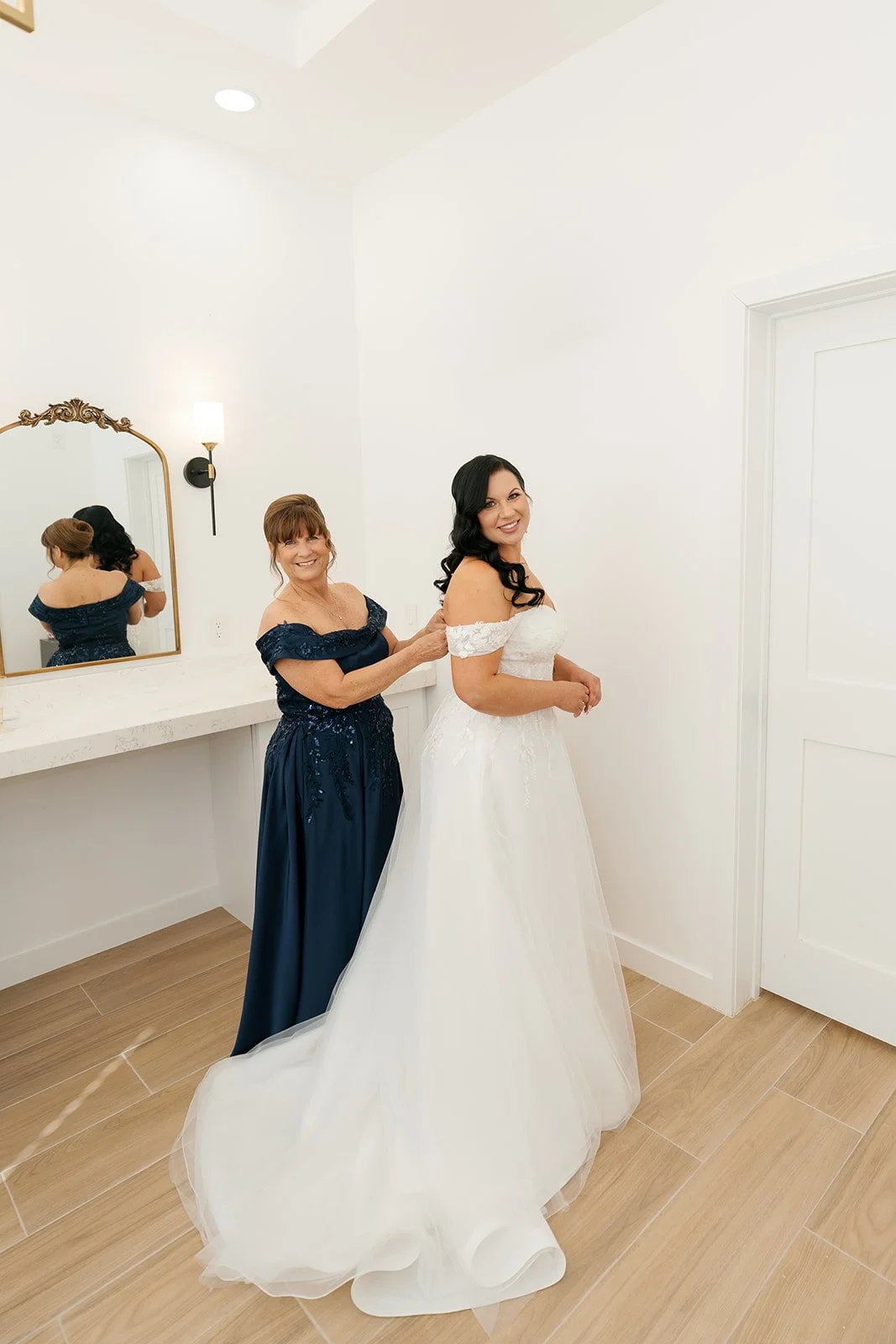 A bride in a white wedding dress smiling as an older woman helps her prepare in a bright, minimalistic room with a large mirror and wooden floor.