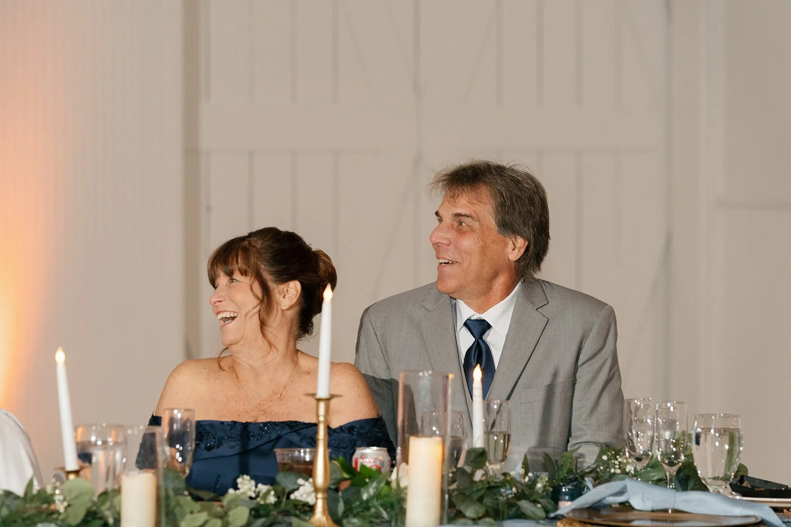 A middle-aged woman and man are sitting at a decorated dining table, both smiling and laughing. The woman is wearing an off-the-shoulder dark blue dress, and the man is in a gray suit with a white shirt and dark blue tie. The table has candles, glass