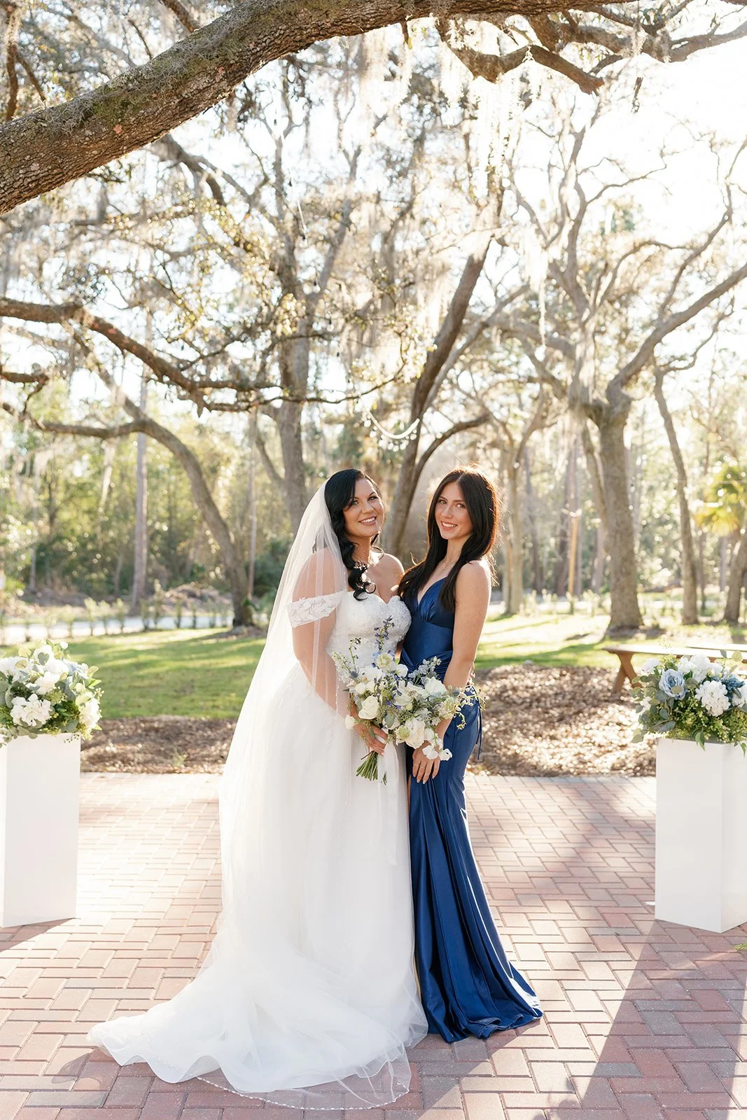 Two women, one in a white wedding dress with a veil and the other in a blue gown, standing outdoors on a brick pathway, holding bouquets of white and purple flowers, on a sunny day with trees in the background.