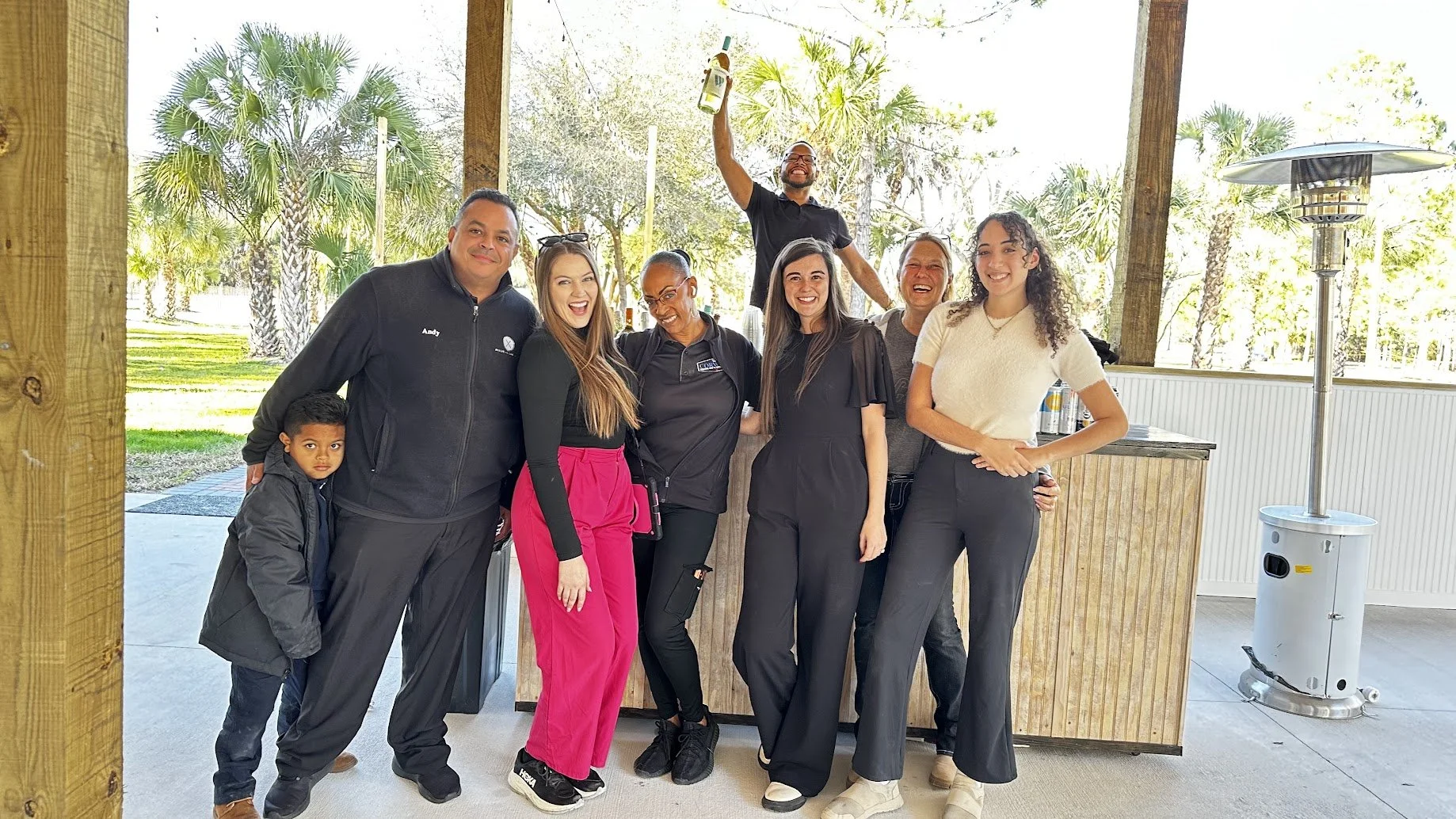 A group of eight people, including a young boy, standing outside near a wooden structure, smiling and posing for the camera during daytime with trees and outdoor heaters in the background.