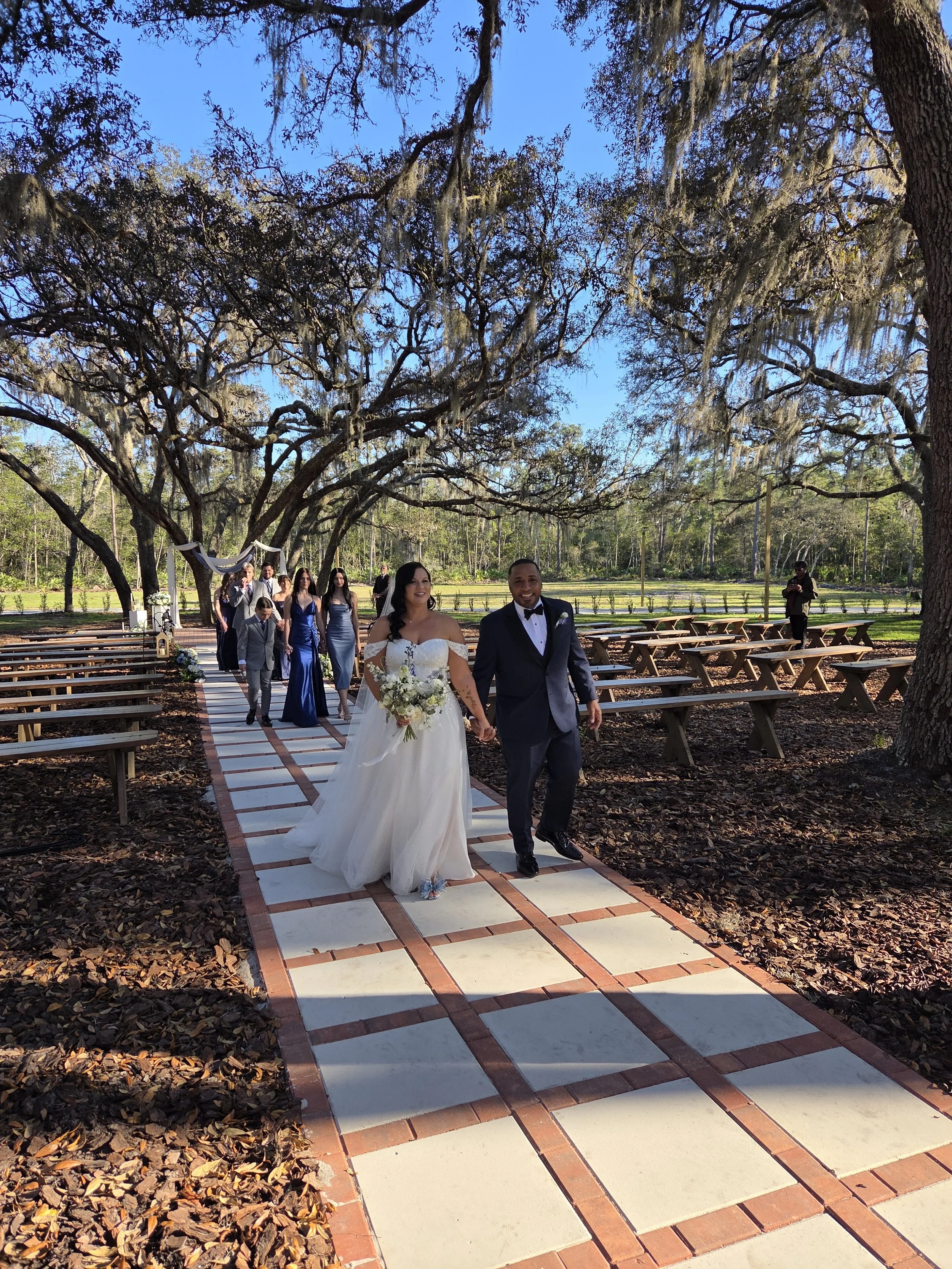 A wedding ceremony outdoors under large oak trees with Spanish moss, featuring a bride in a white gown and a groom in a tuxedo, walking down a brick and concrete aisle, followed by bridesmaids and groomsmen, with a person taking photos in the backgro