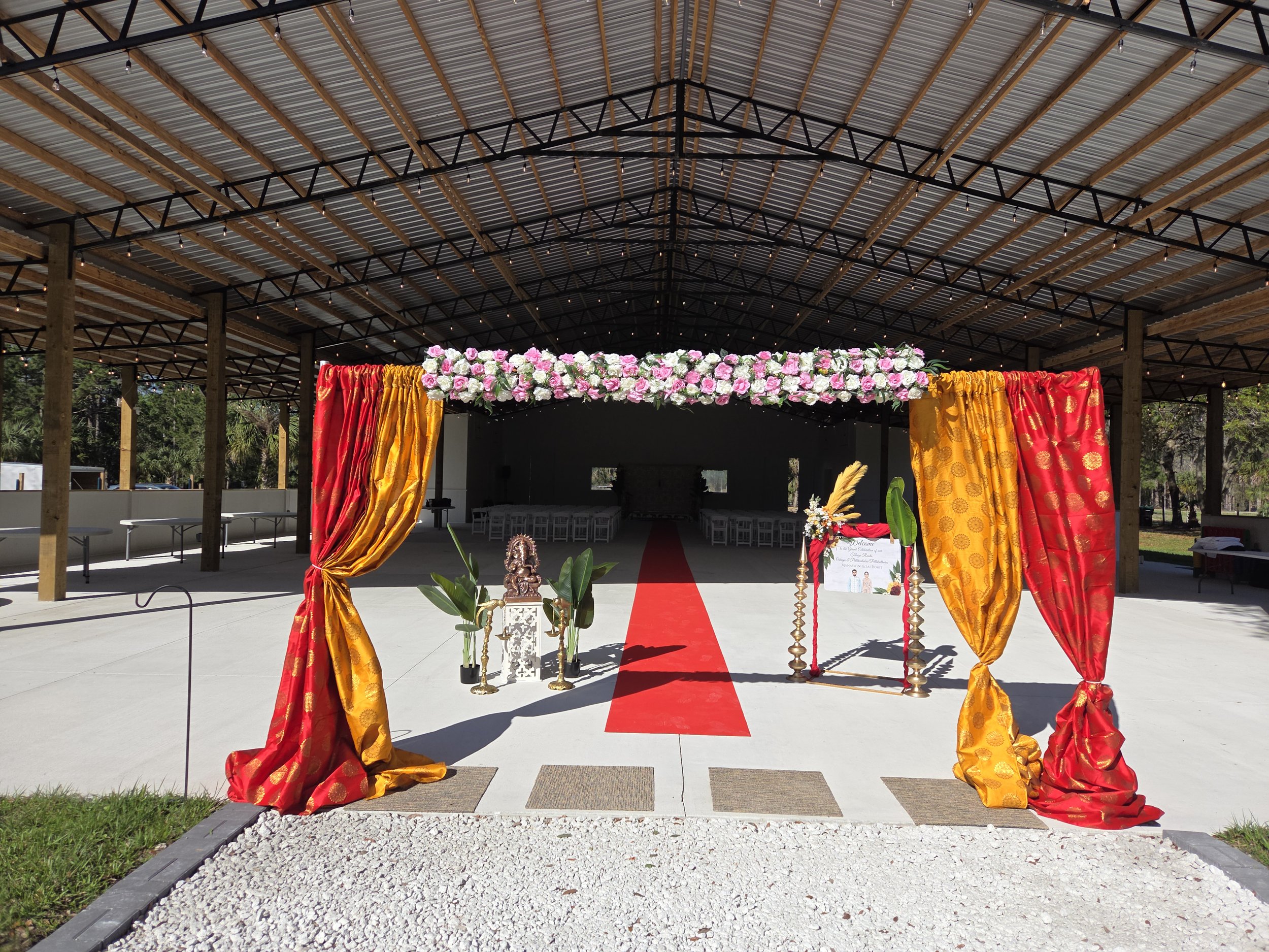 Outdoor wedding ceremony setup with a red carpet leading to a decorated entrance with red and yellow curtains, and a floral arrangement of pink and white flowers underneath a covered open-air structure.