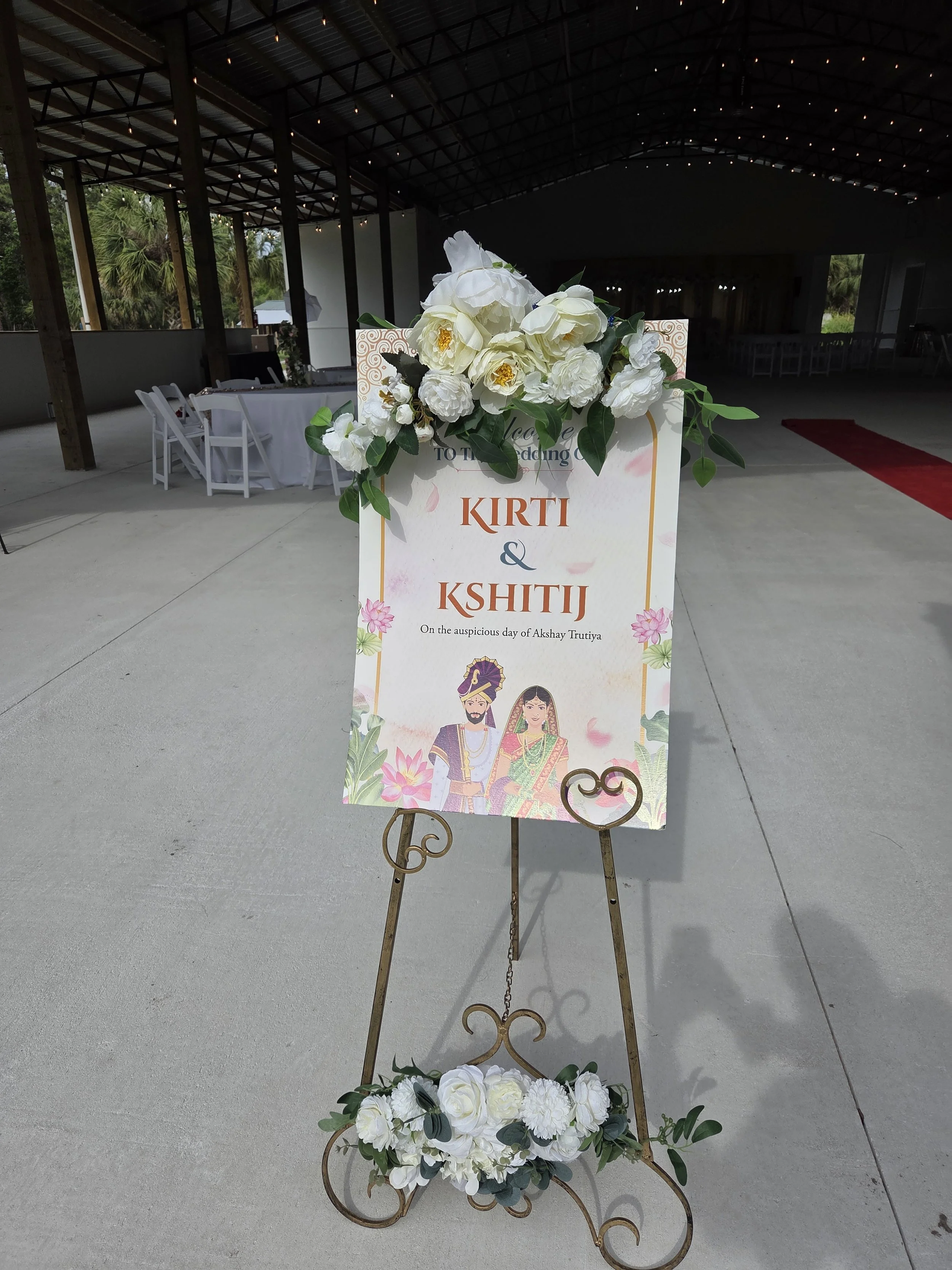 A wedding sign with white flowers and green leaves, featuring images of a bride and groom in traditional Indian attire, and the names 'Kirti and Kshitij' with a message about the auspicious day.