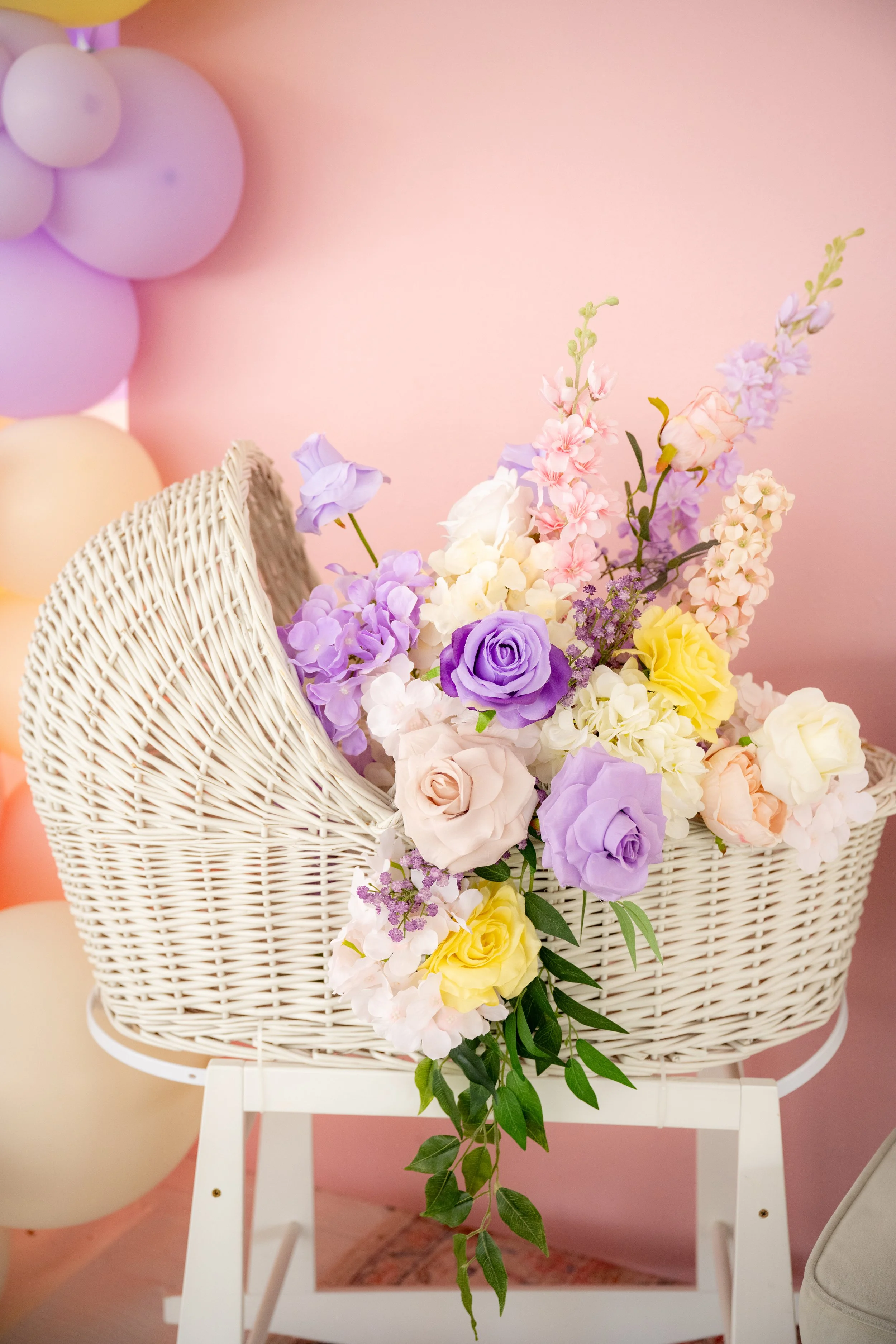 A wicker basket filled with pastel-colored flowers, including roses and peonies, on a white stand against a pink wall with balloons in the background.