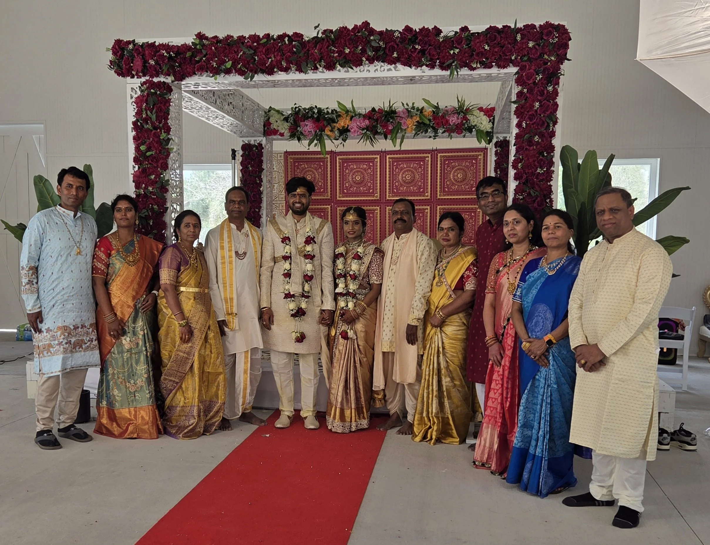 A group of people dressed in traditional Indian attire standing under a decorated floral canopy at a wedding ceremony. The bride and groom are in the center, with the bride wearing a cream and gold saree and the groom in white and gold, both wearing 
