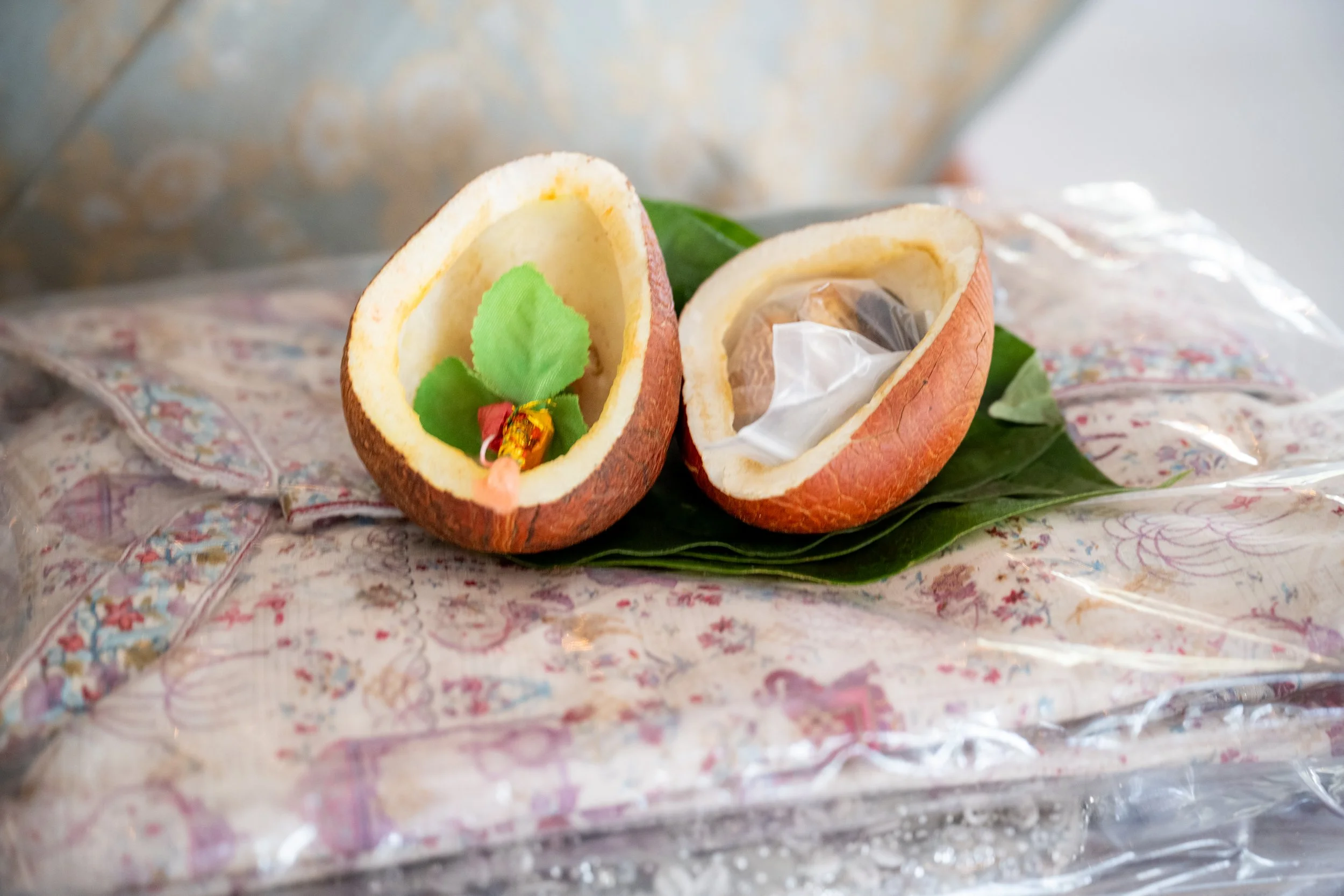 Cut open coconut shell with a green leaf inside, containing small decorative items, placed on a leaf and decorative paper.