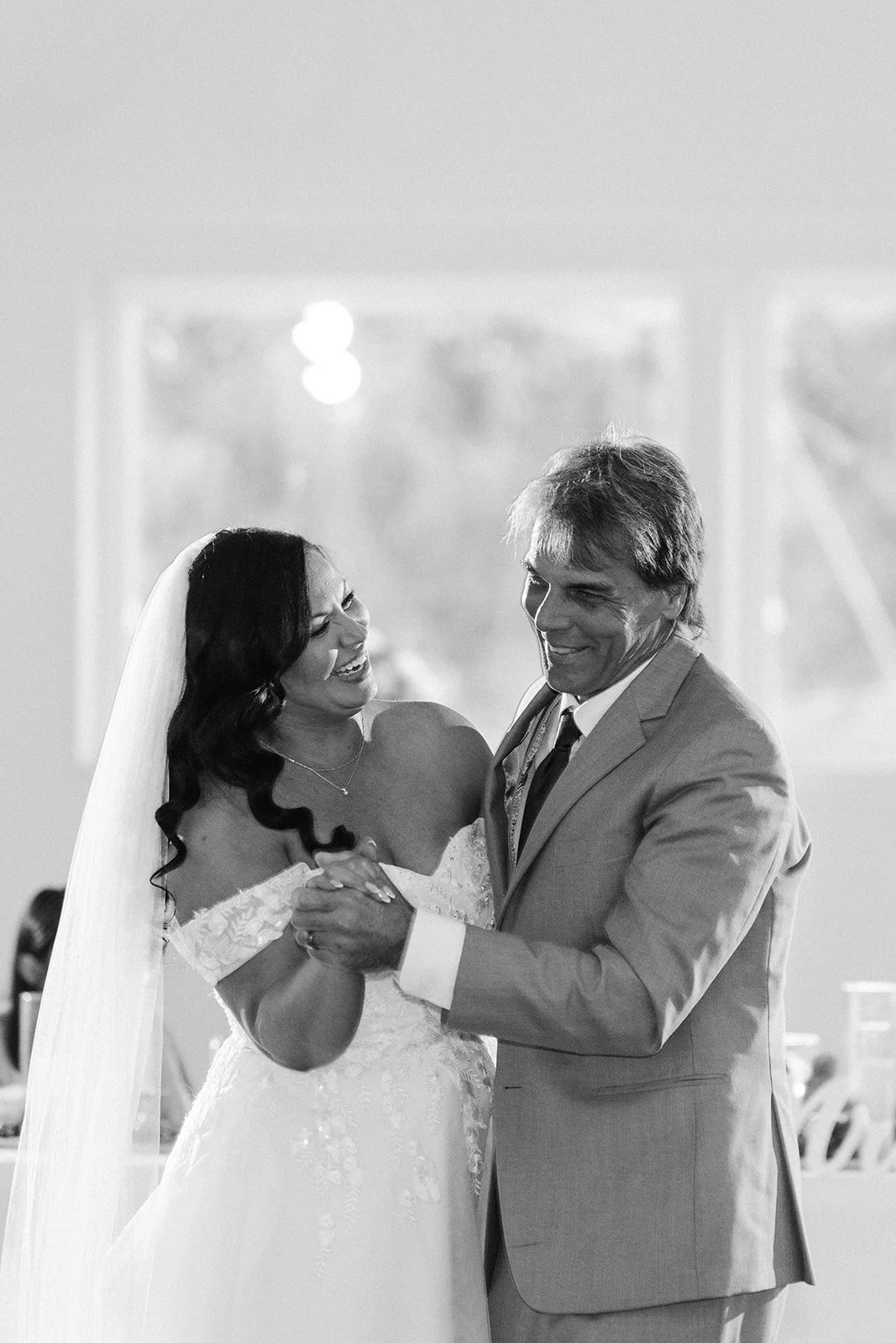 A bride and an older man, likely her father, dancing together at a wedding reception, both smiling and holding hands, in black and white.