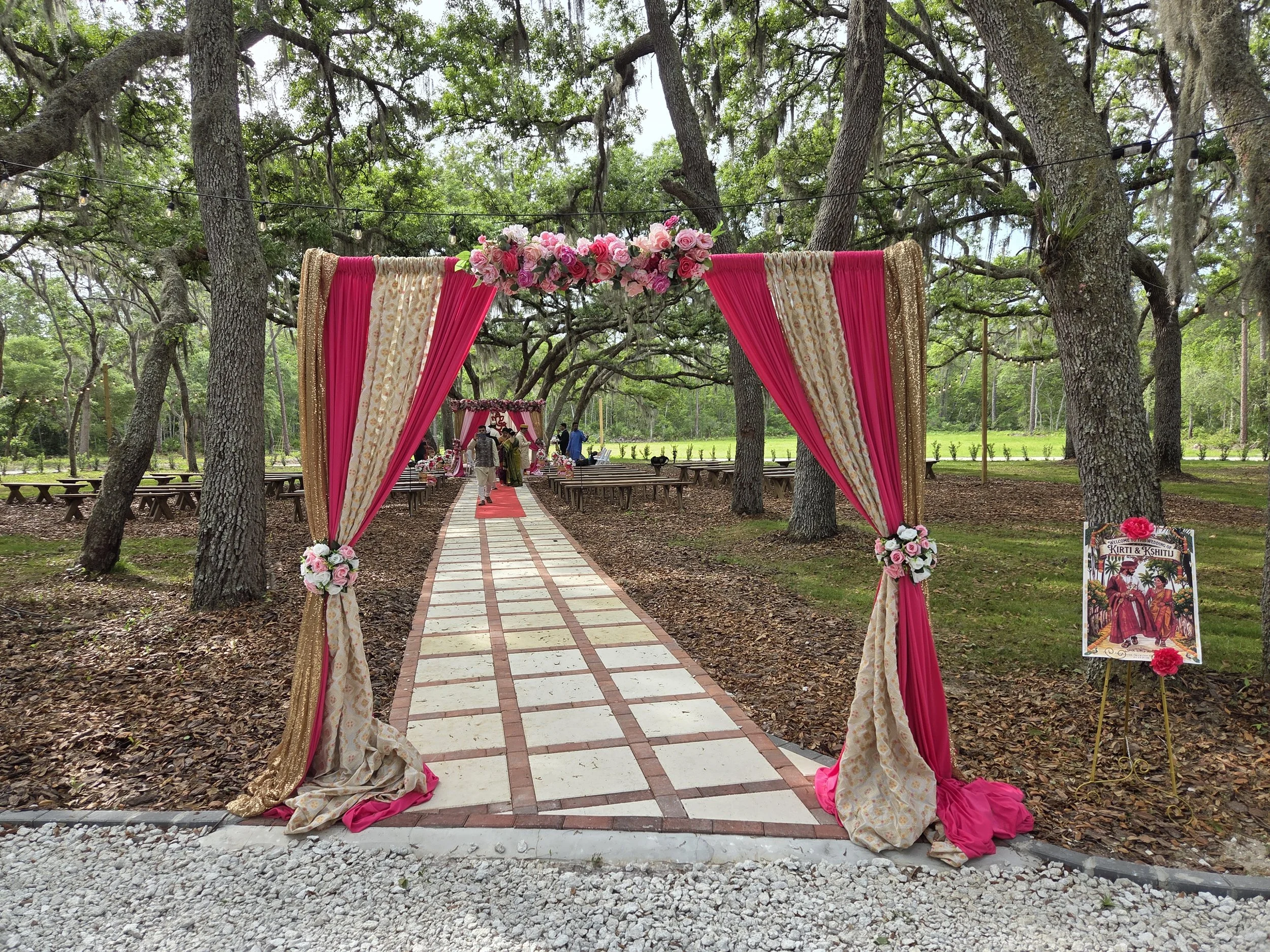 An outdoor wedding setup featuring a pink and gold decorated archway with floral arrangements, set on a pathway through a wooded area. There are benches and guests visible in the background.