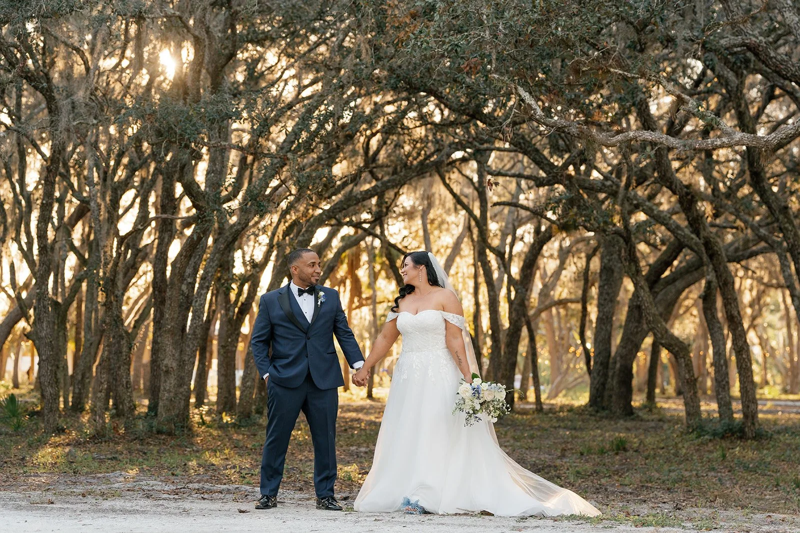 A bride and groom standing outdoors in a wooded area during sunset, holding hands and smiling at each other. The bride wears a white off-shoulder wedding gown and holds a bouquet, while the groom is dressed in a navy blue tuxedo with a black bow tie.