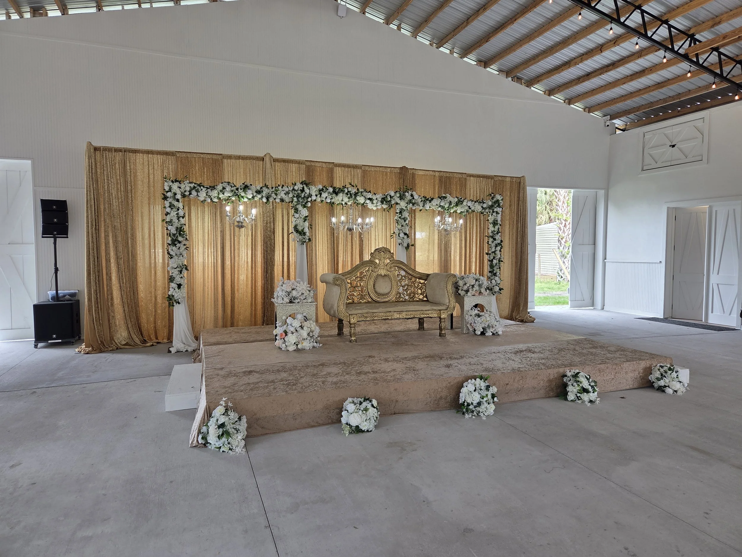 Indoor wedding stage decorated with floral arch, gold curtains, and elegant sofa.