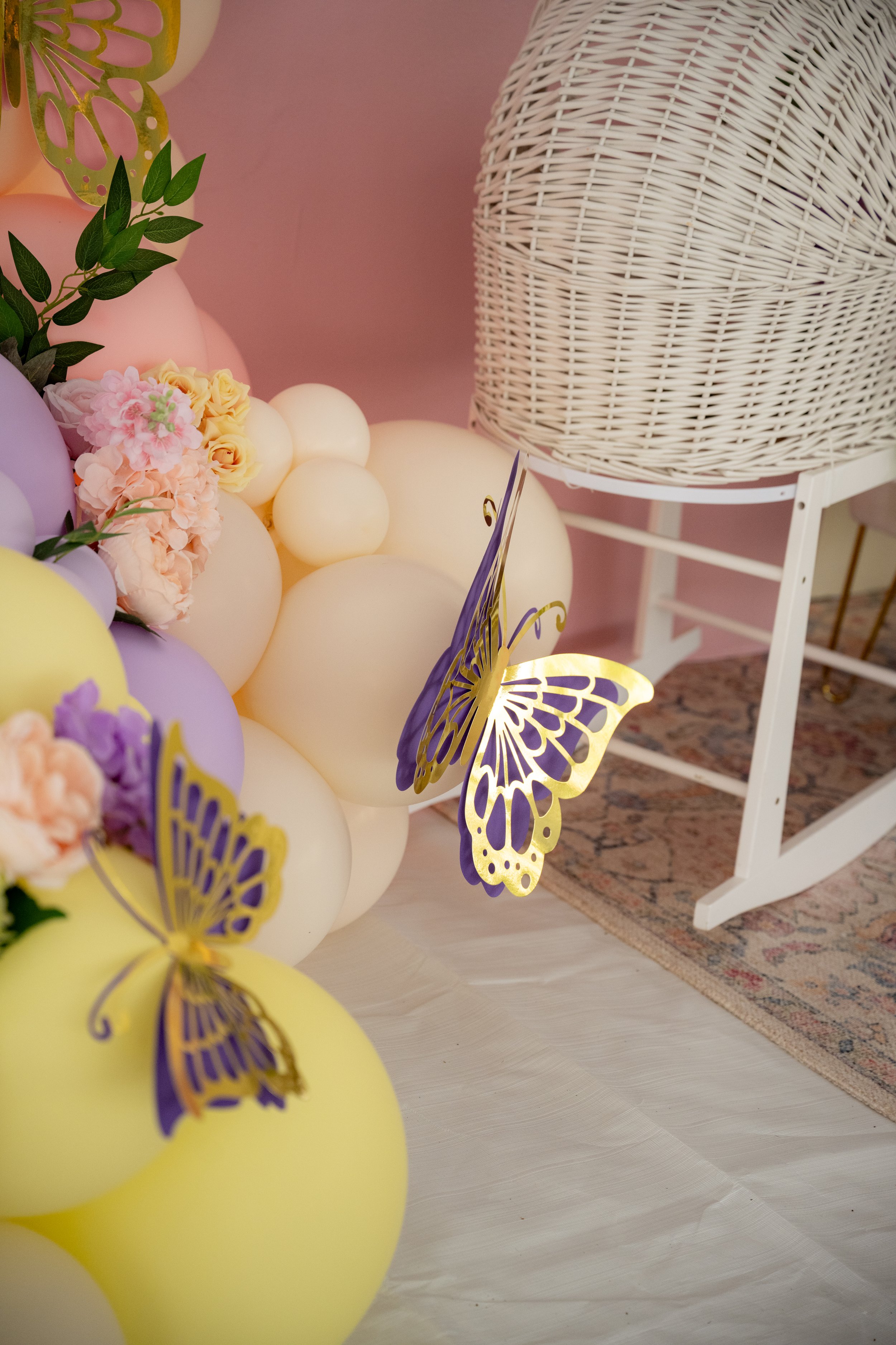Colorful balloons with flowers and butterfly decorations arranged on a wall near a white wicker chair and rug.