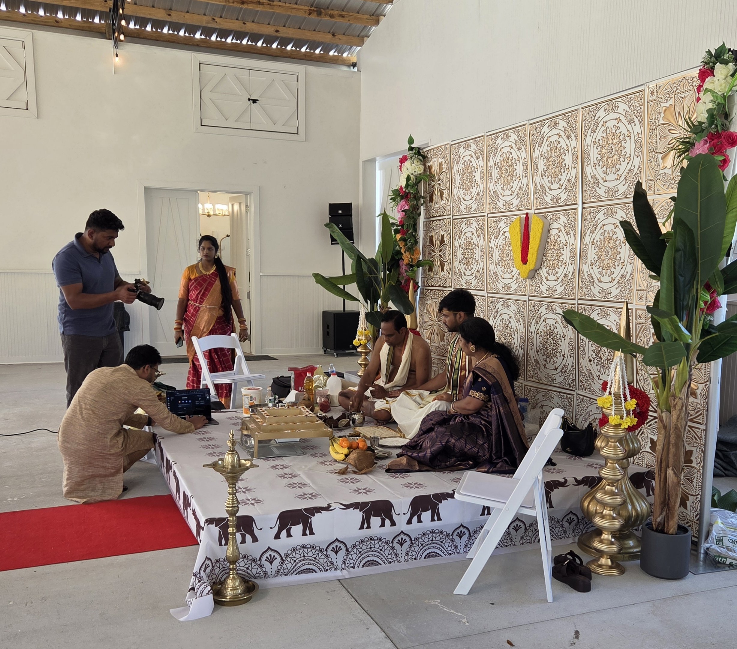A wedding or cultural ceremony setup with a decorated table, floral arrangements, and traditional clothing. People are gathered, some sitting at the table, others standing nearby.